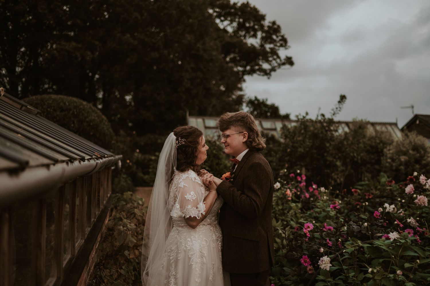 A romantic wedding couple embraces in front of a rustic greenhouse surrounded by blooming pink flowers in moody lighting.