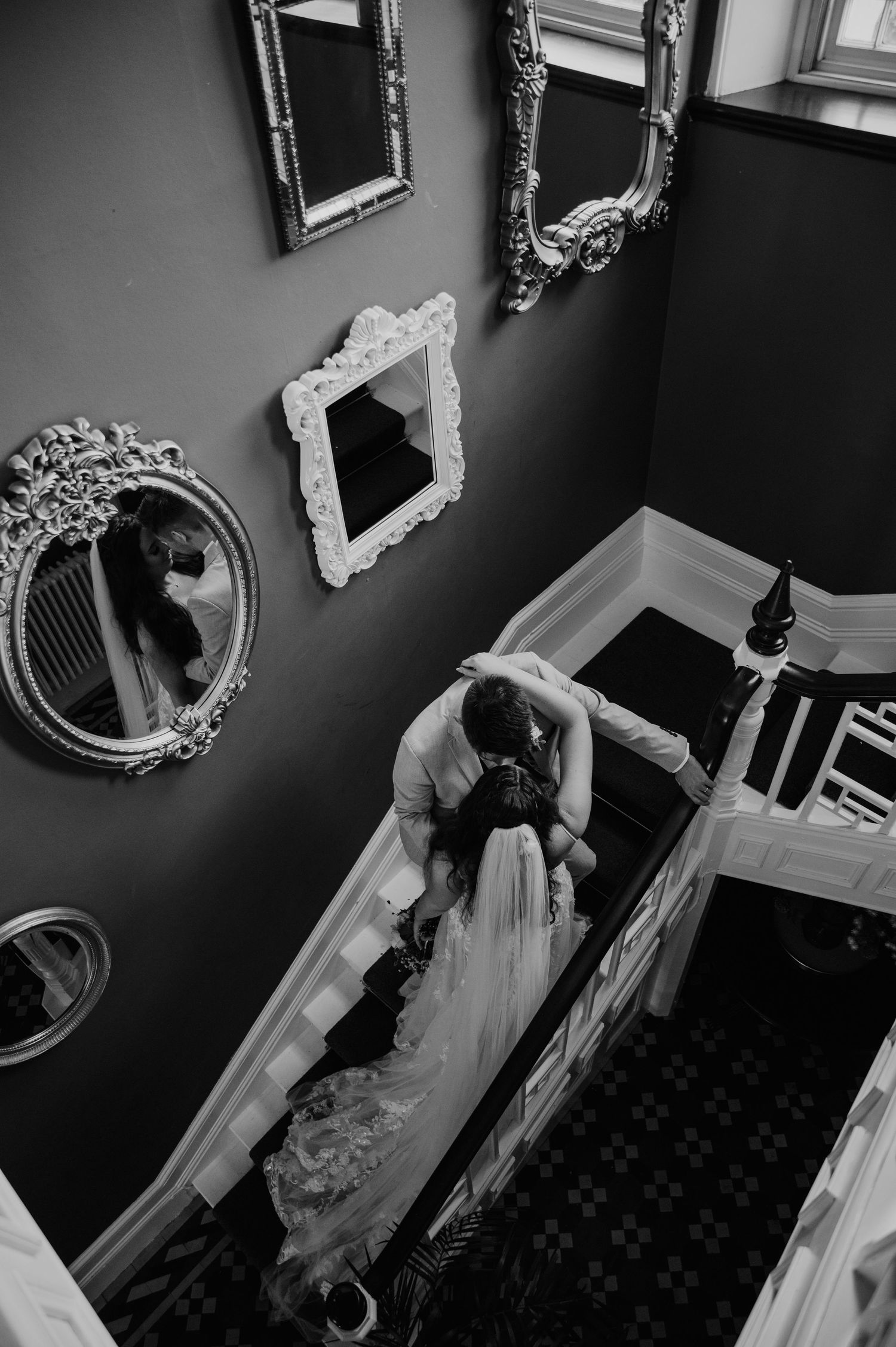 Black and white view from above of ornate vintage mirrors on dark walls and a white staircase in a home interior at Beaconside House.