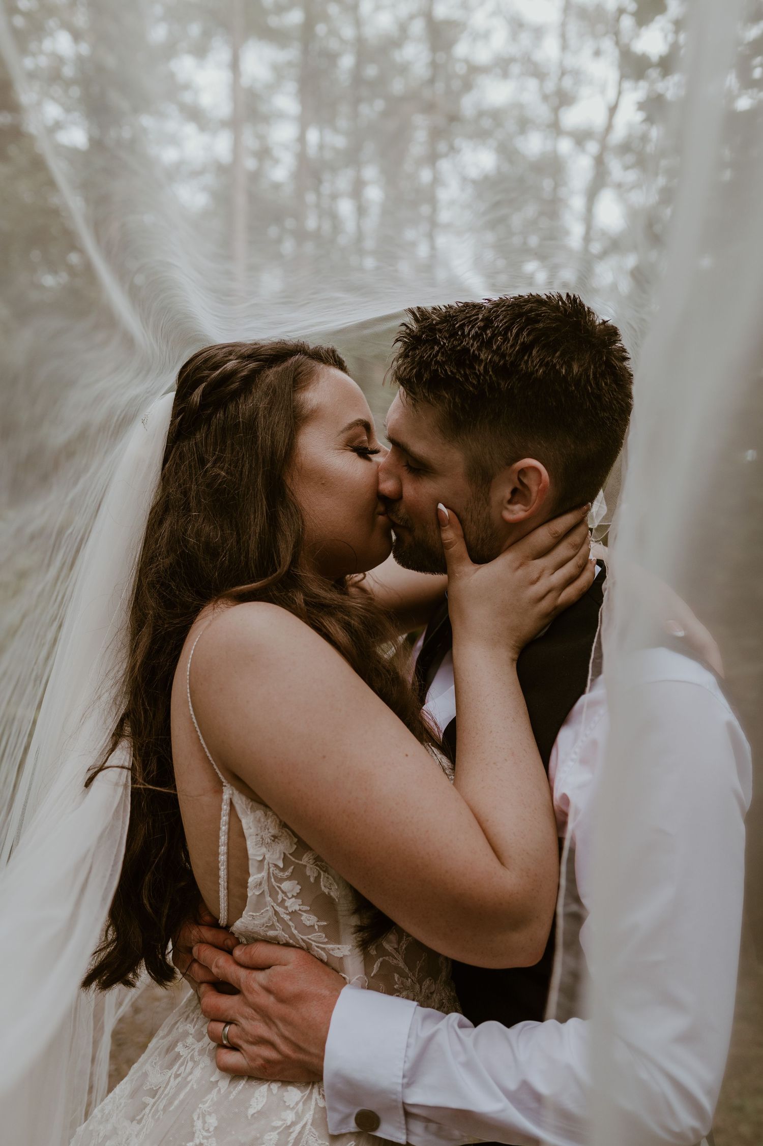 A wedding couple shares an intimate kiss in a misty forest setting while the bride's veil flows behind them.