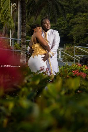 Romantic couple embrace in tropical garden setting wearing white and gold formal attire.