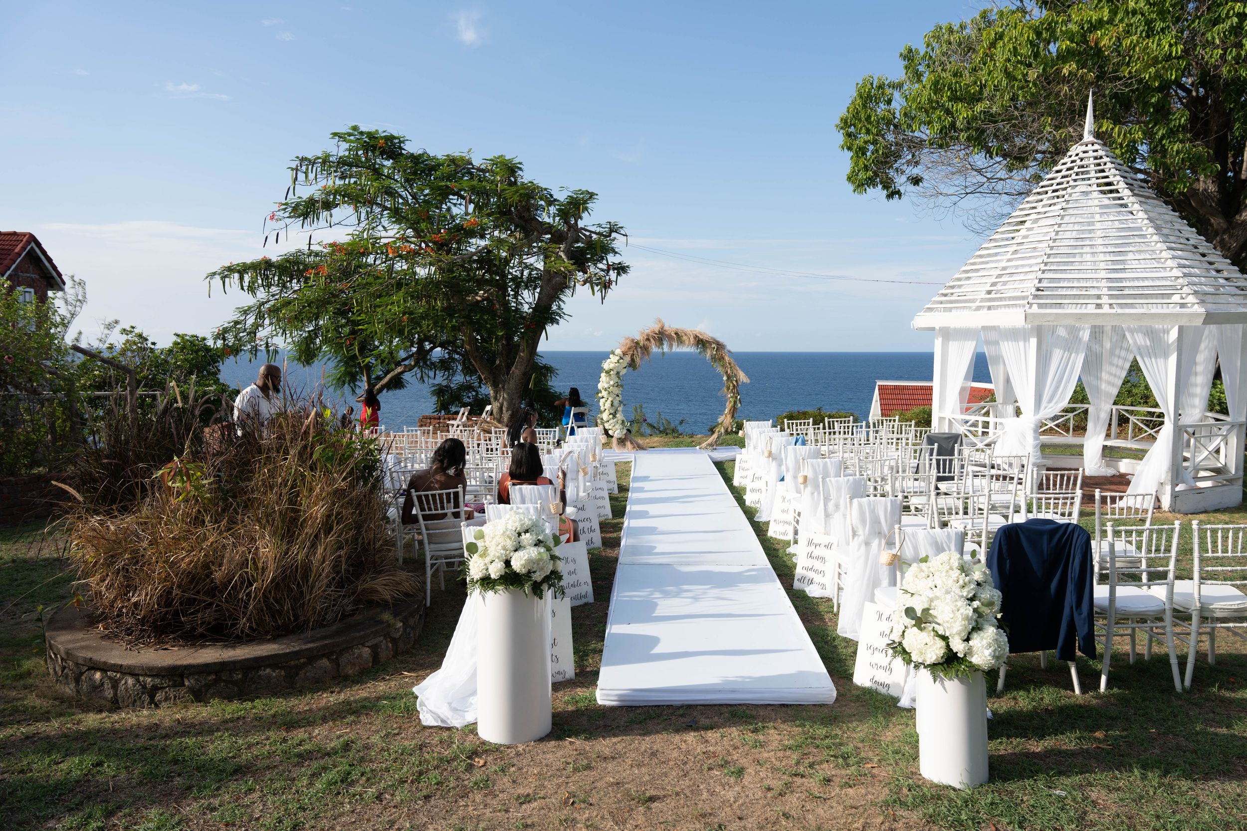 White wedding ceremony setup with gazebo and ocean view featuring white chairs and floral arch.