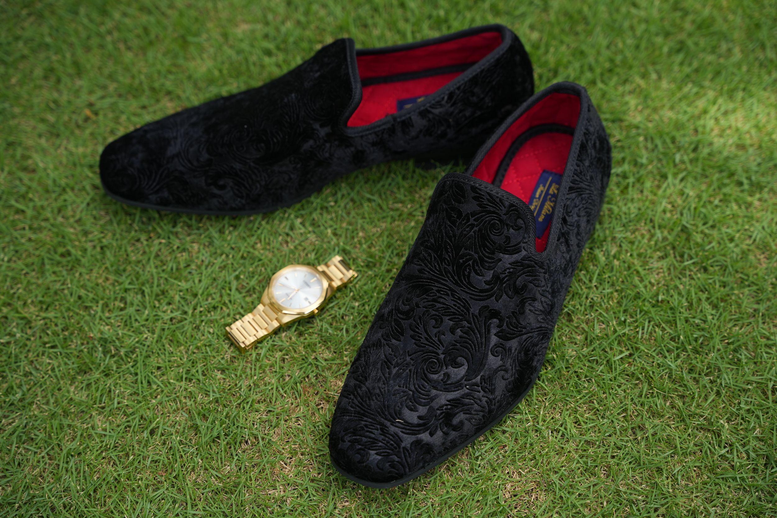 Black velvet loafers and gold watch displayed on green grass.