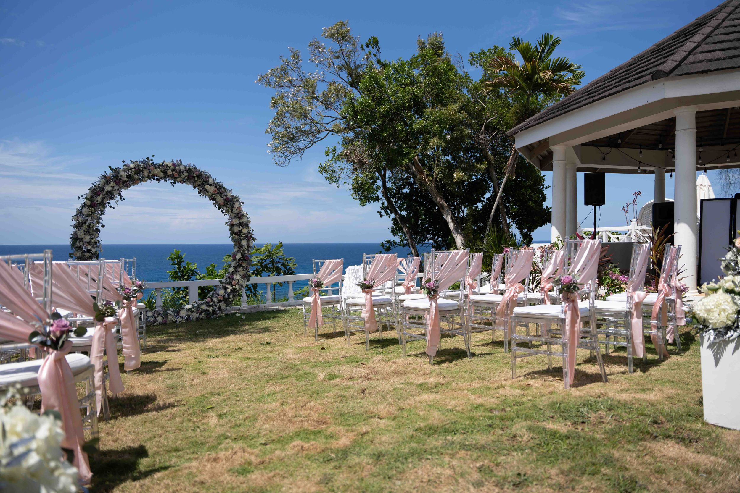 Wedding ceremony setup with white chairs and floral arch overlooking the ocean on a sunny day.