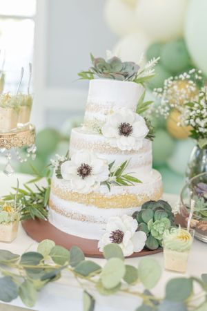 White tiered cake decorated with succulents, white flowers, and gold accents on a dessert table.