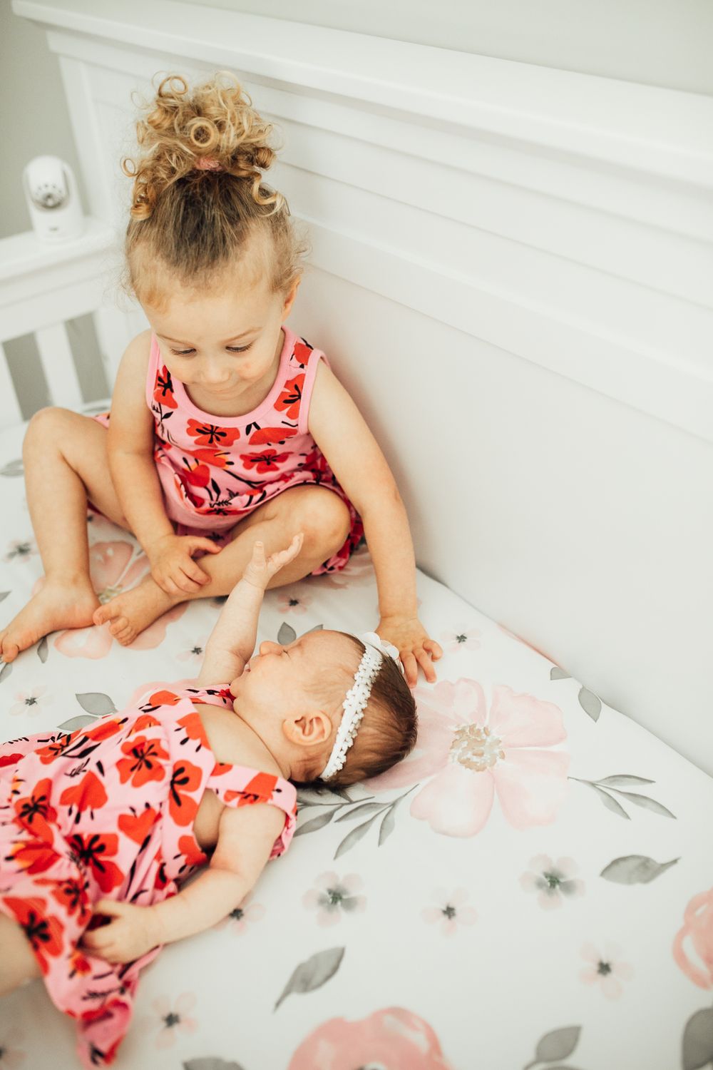 Two toddlers in matching floral red dresses sit together on a white bed with floral print sheets.