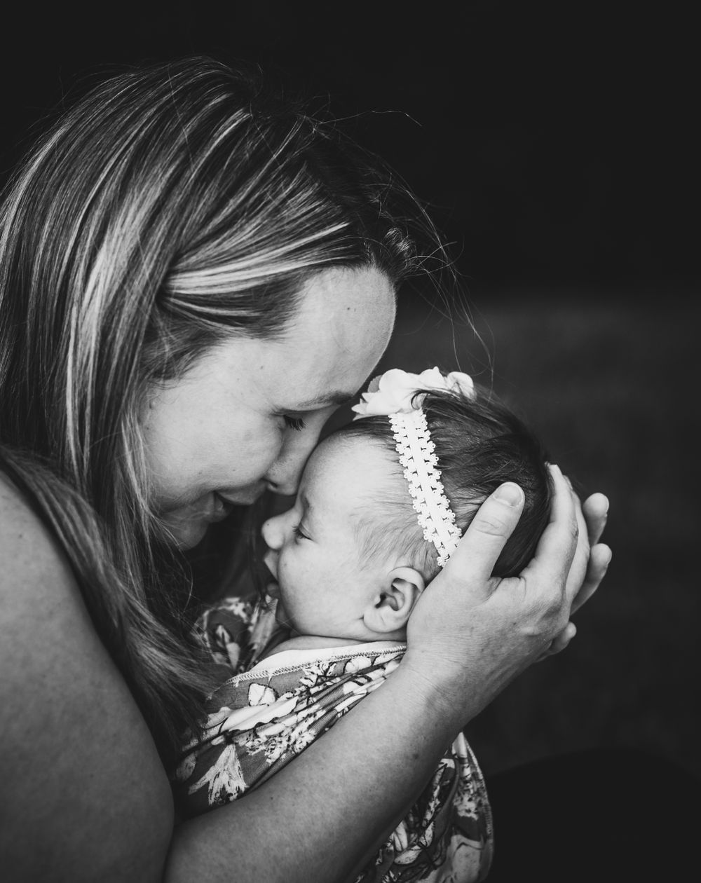 Black and white intimate portrait showing tender moment between parent and infant wearing floral outfit and white headband.
