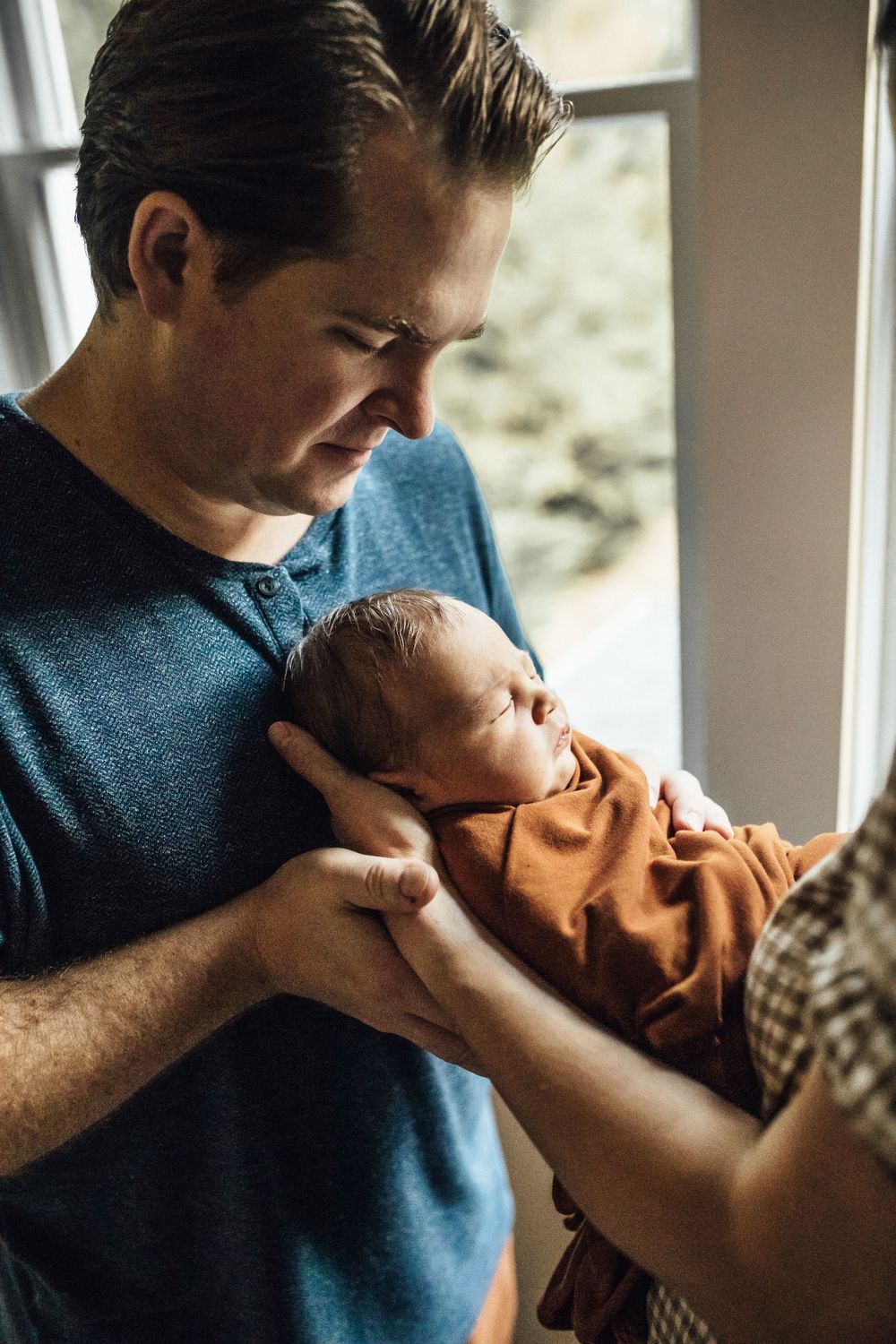 A parent in a blue shirt tenderly holds a newborn baby wrapped in a brown swaddle by a window in natural light.