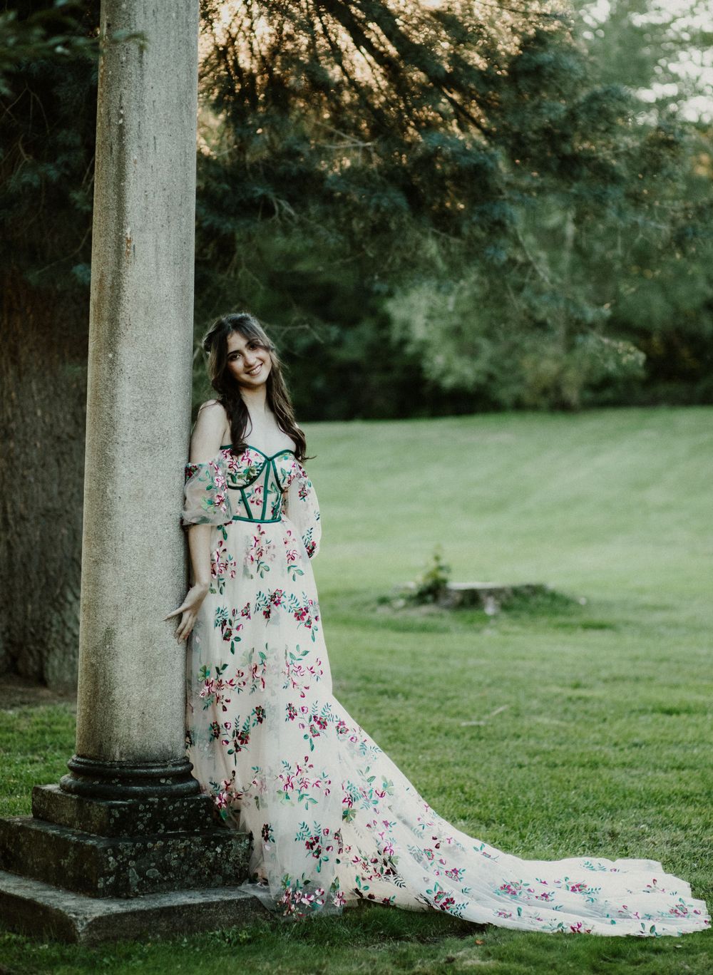 Person in flowing floral dress poses elegantly beside classical stone pillar in garden setting.