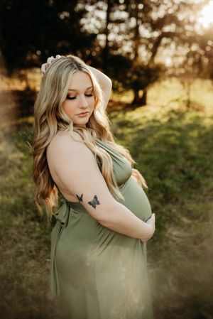 Pregnant woman in olive green maternity gown with butterfly tattoo posing in golden sunset light.