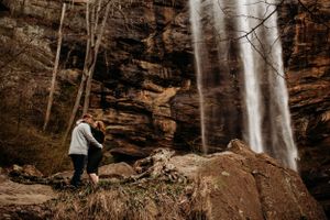 Couple holding hands near a stunning waterfall cascading over rocky cliffs in the woods.