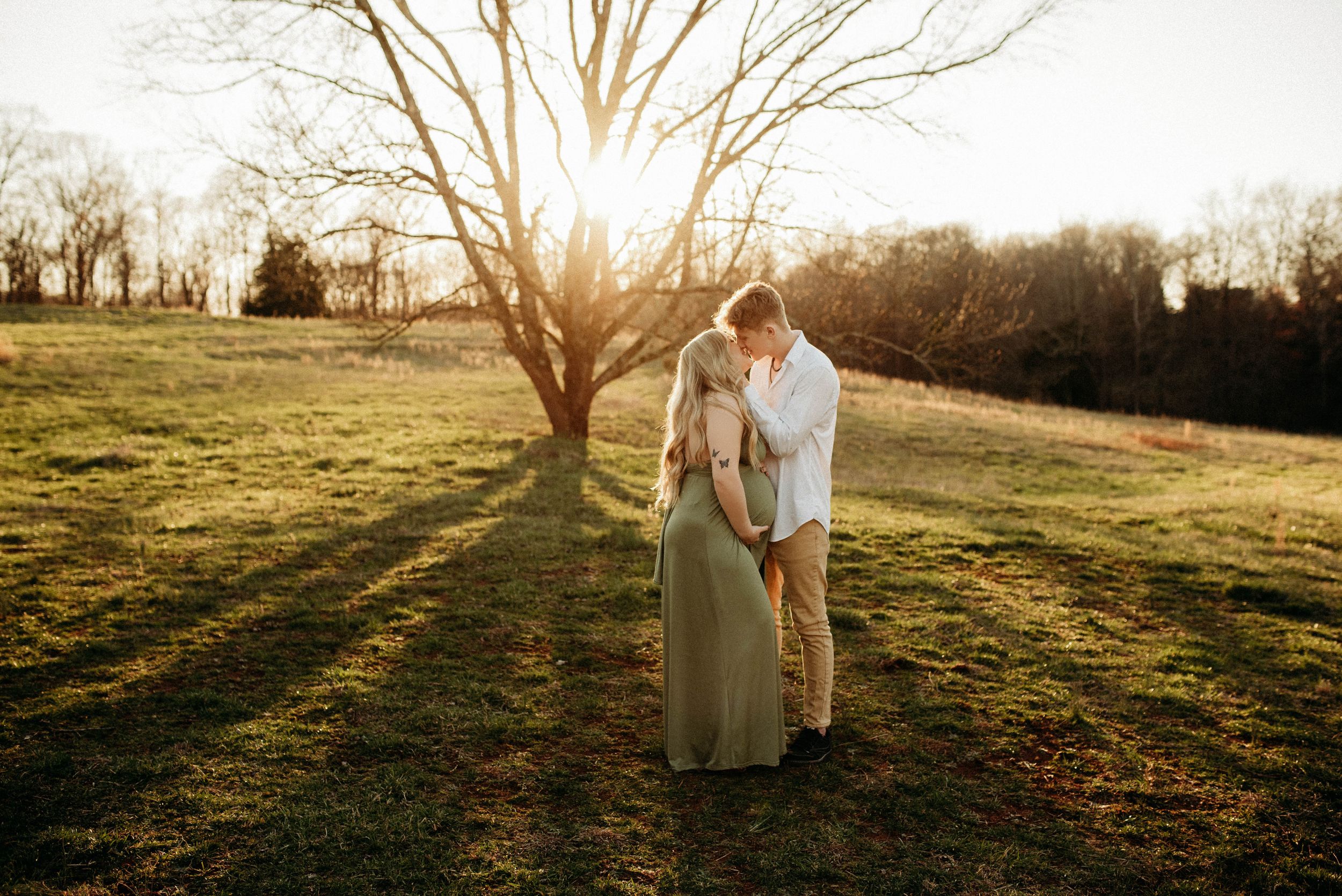 Couple sharing a kiss in a sunlit field during a maternity photo session at golden hour.
