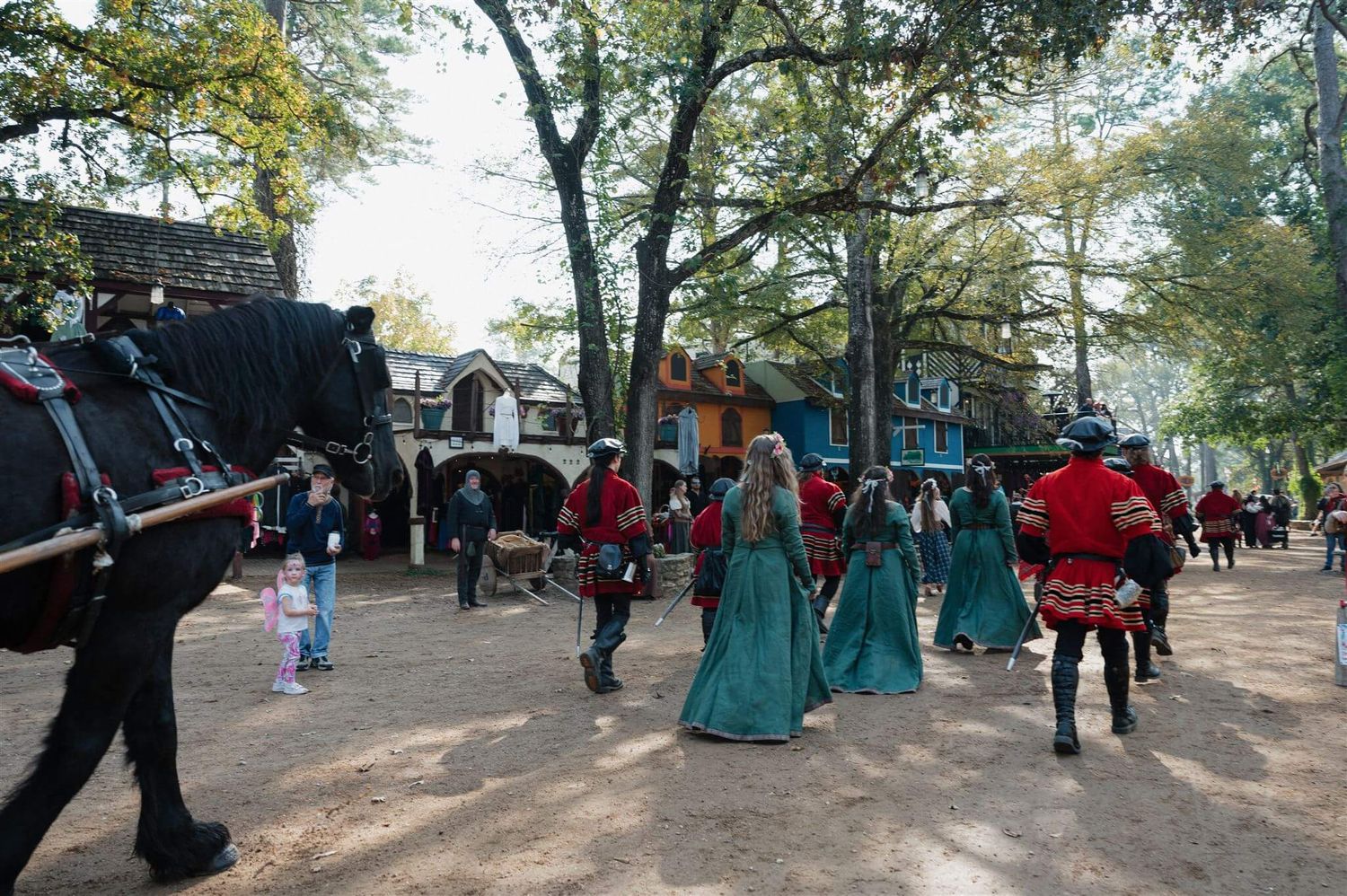 Medieval costumed performers and guests walk along a tree-lined path at a renaissance festival.