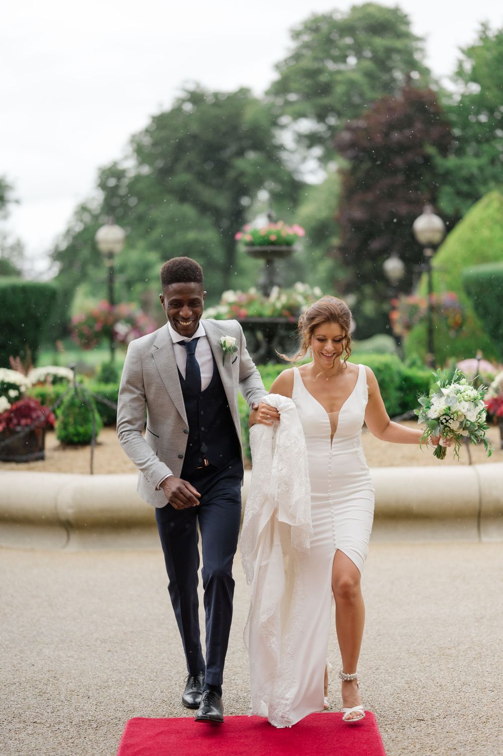 A smiling couple walks down a red carpet in a garden, bride in white dress holding bouquet, groom in gray suit at Killashee Hotel wedding