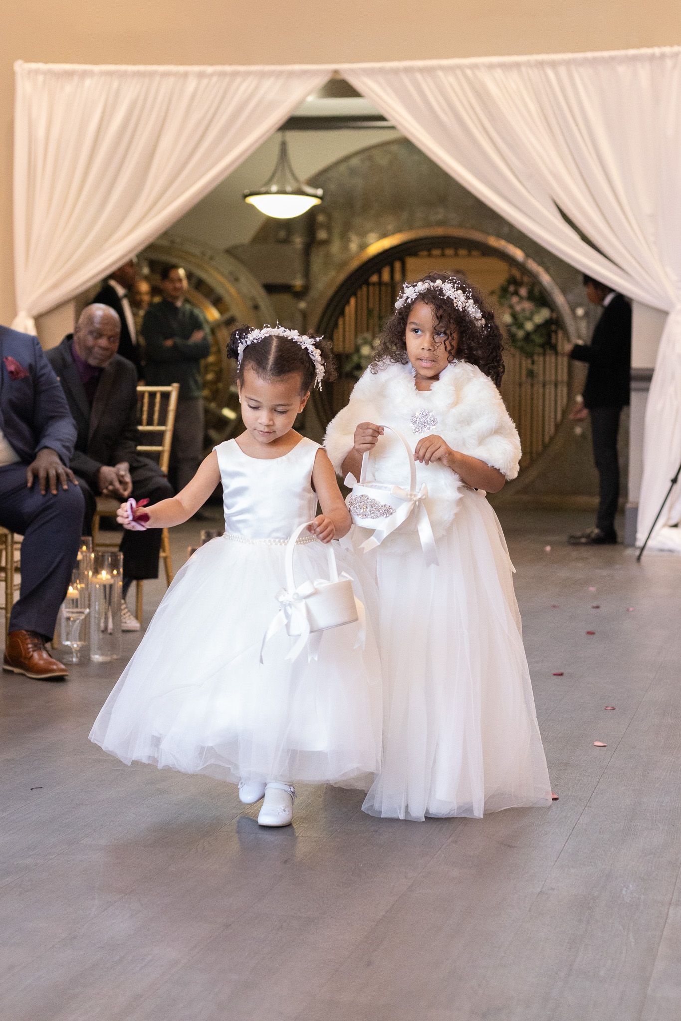 Two young flower girls in white dresses walking down aisle at wedding ceremony.