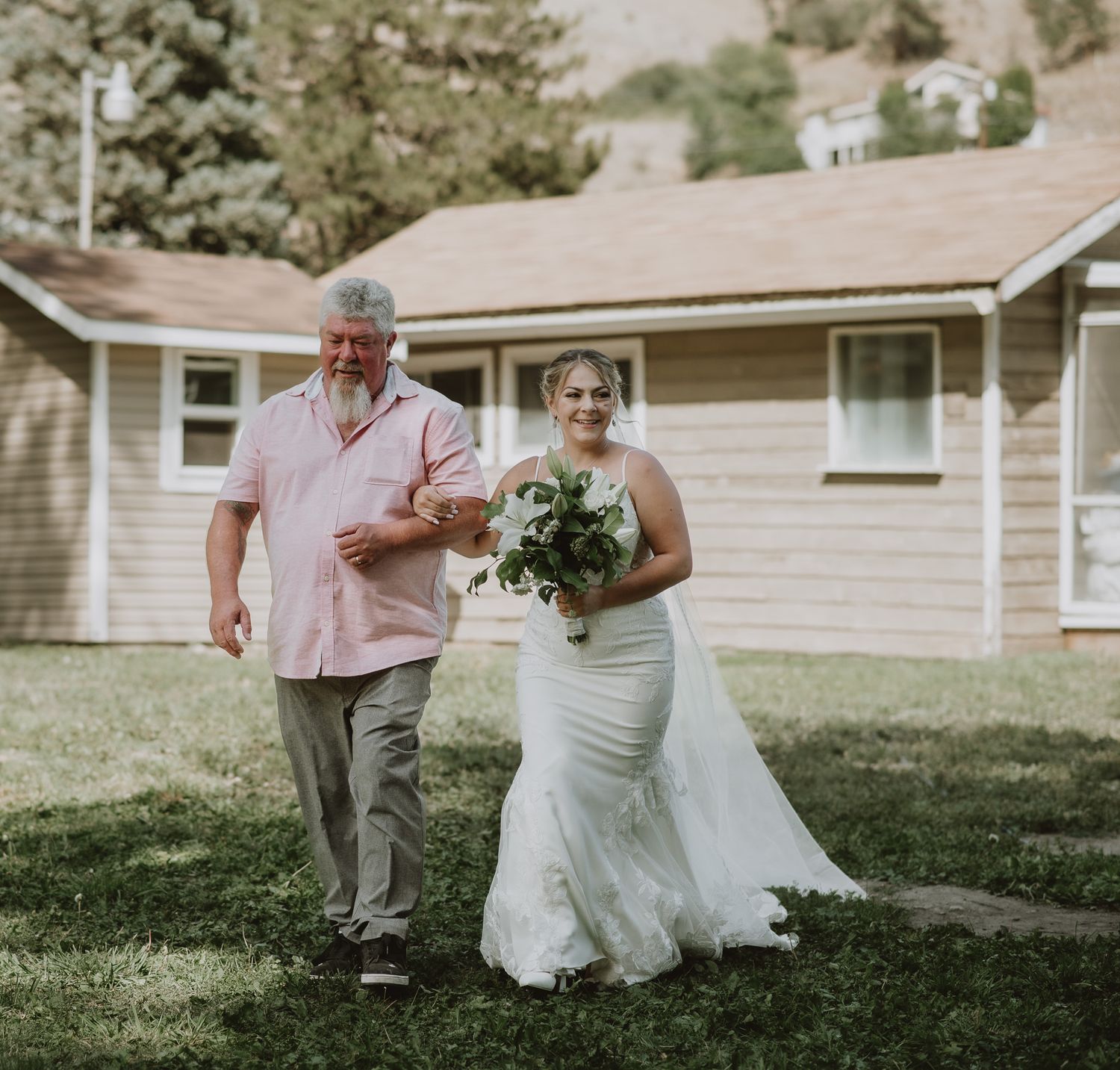 The bride being walked down the aisle by her dad. She is holding her bouquet and smiling in Summerland