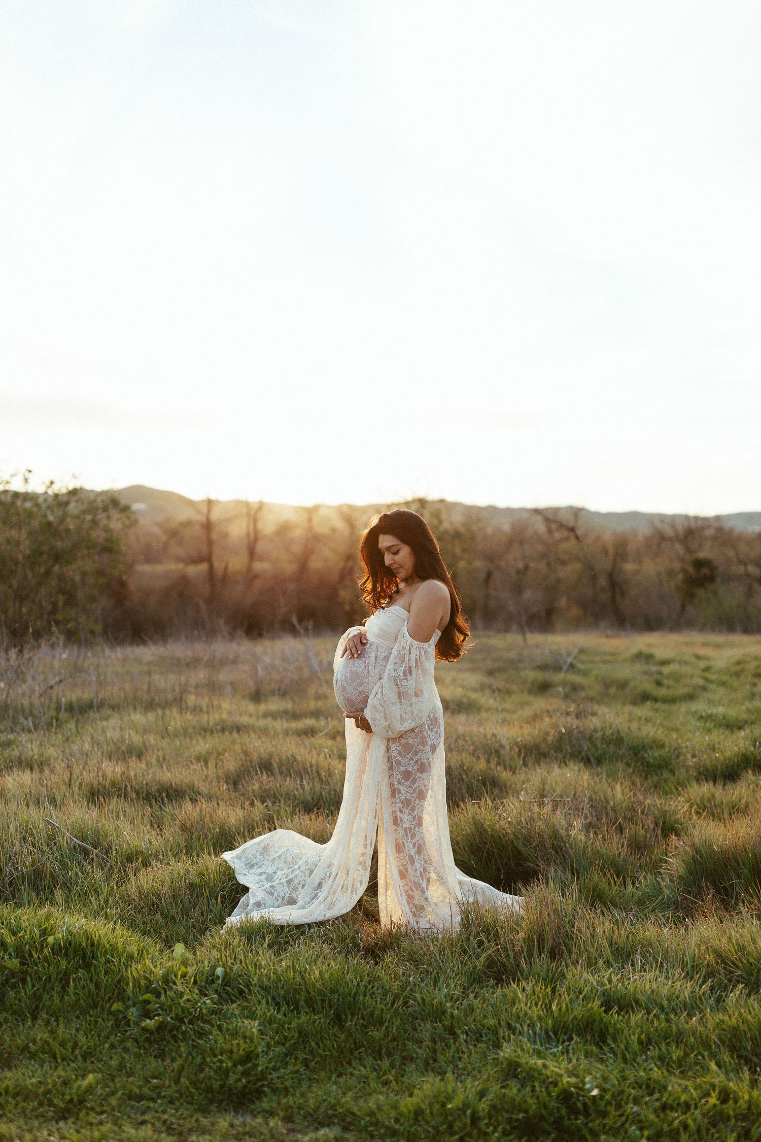 A pregnant woman poses in a white lace gown during golden hour in a grassy field.