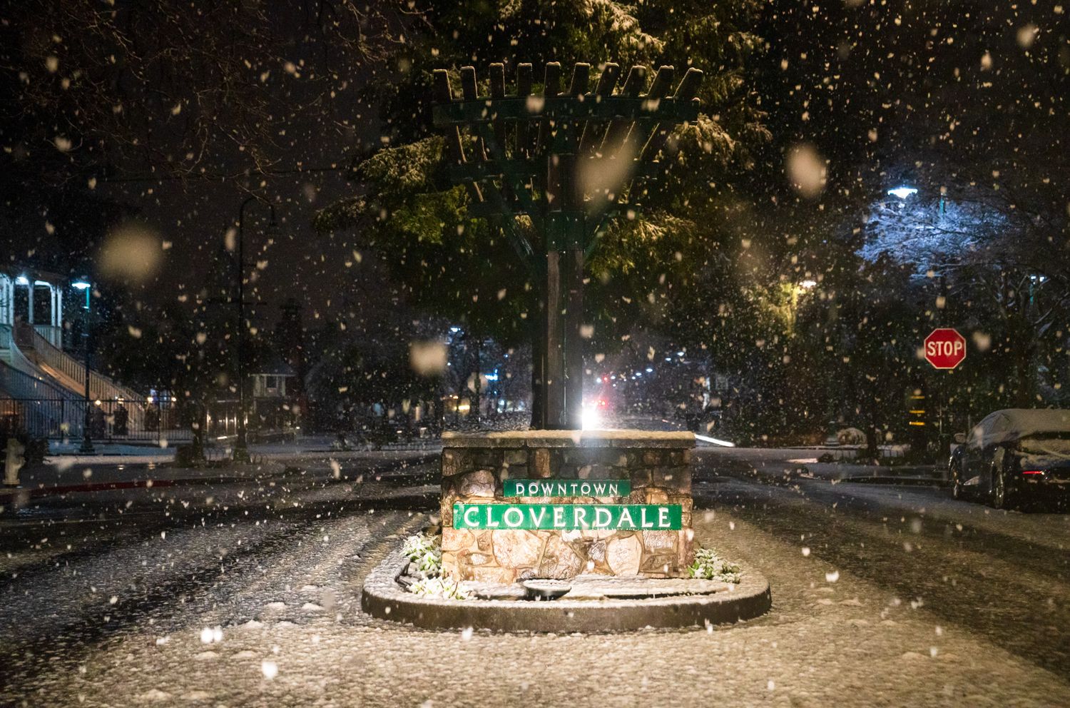 Snowy night scene of an illuminated town entrance sign with falling snowflakes and street lights.
