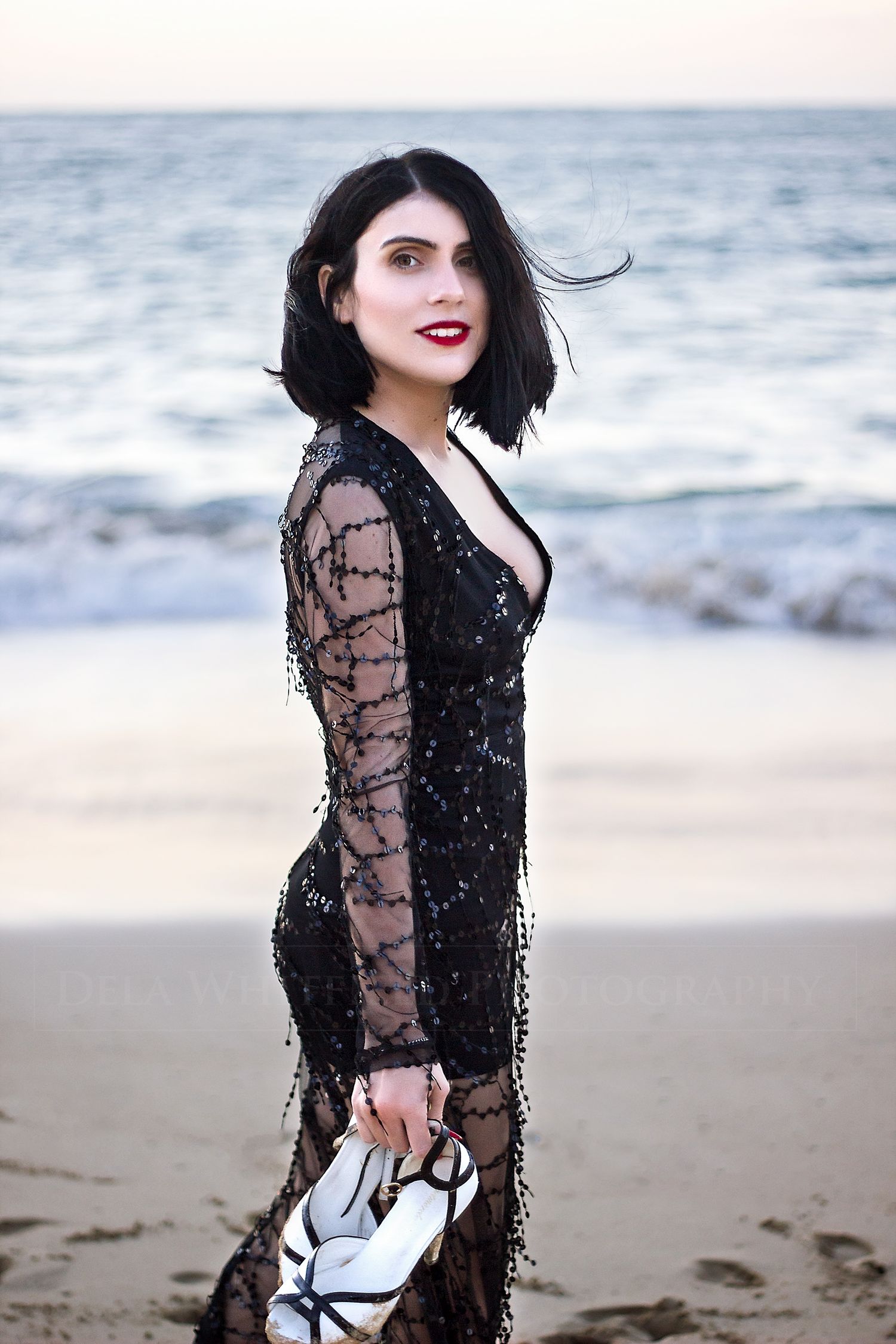 Woman in beaded gown at sunset on beach, carrying her high heel shoes with the ocean in the background.