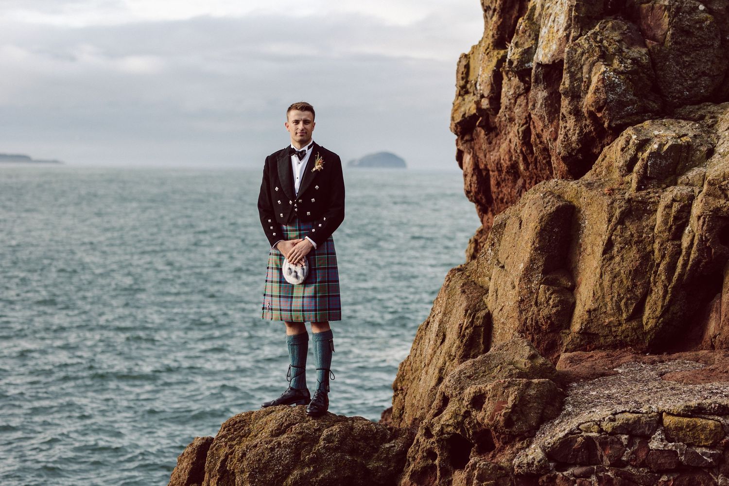 Groom in traditional Scottish kilt stands on rocky coastal cliffs overlooking the sea.