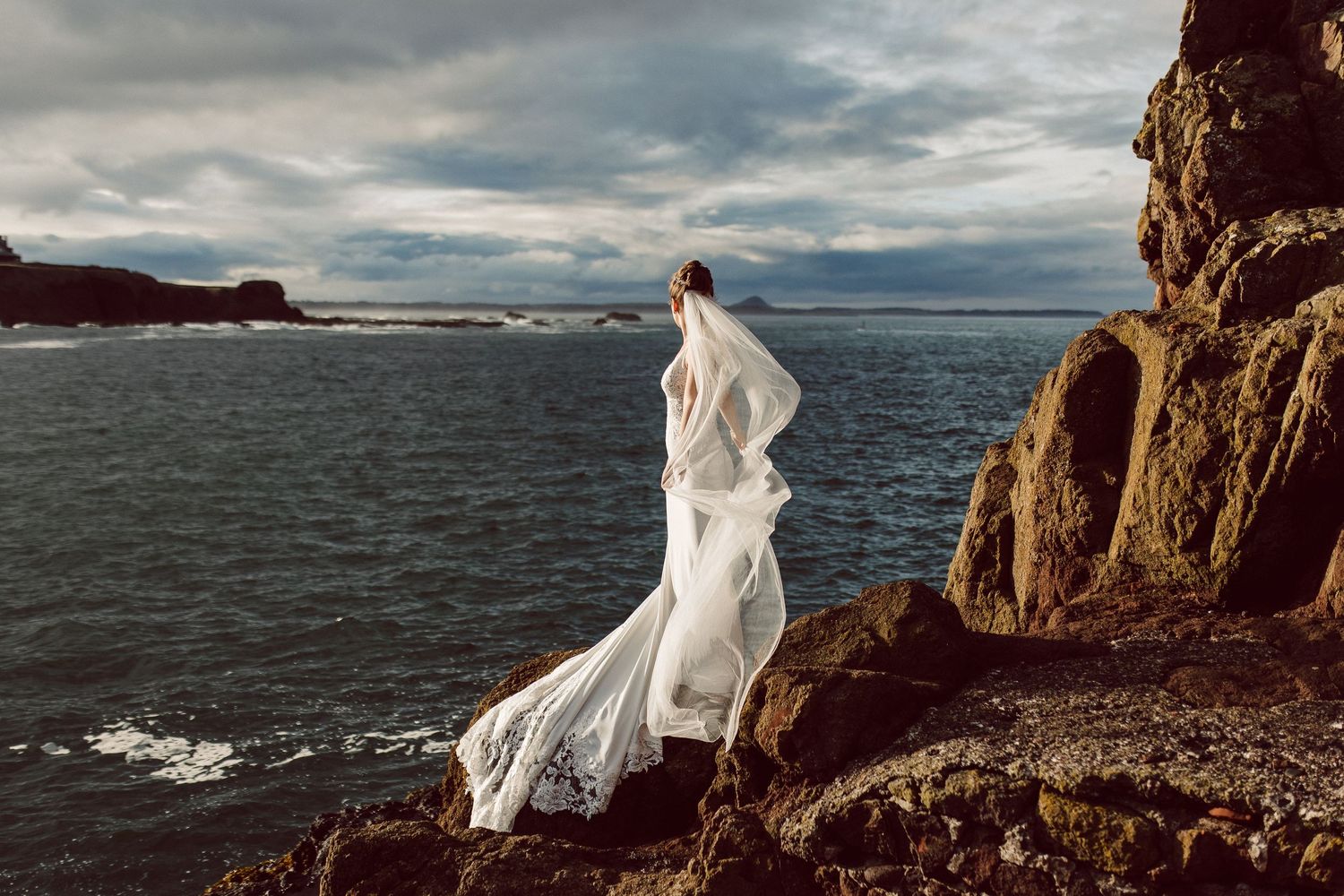 White wedding dress and veil flow dramatically in the wind against coastal cliff backdrop.