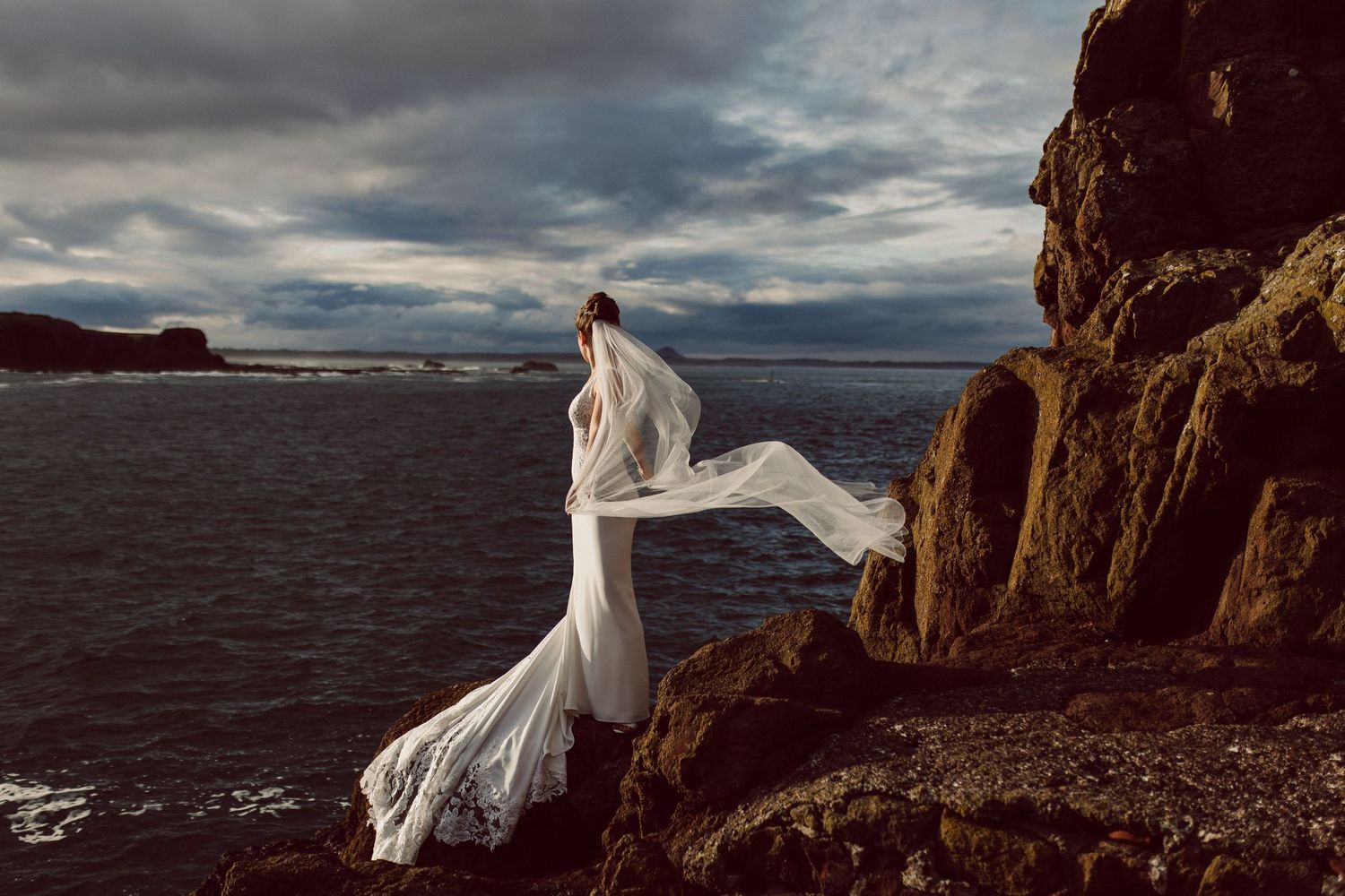 Bride's dress billows in the wind on dramatic clifftop overlooking stormy seas.