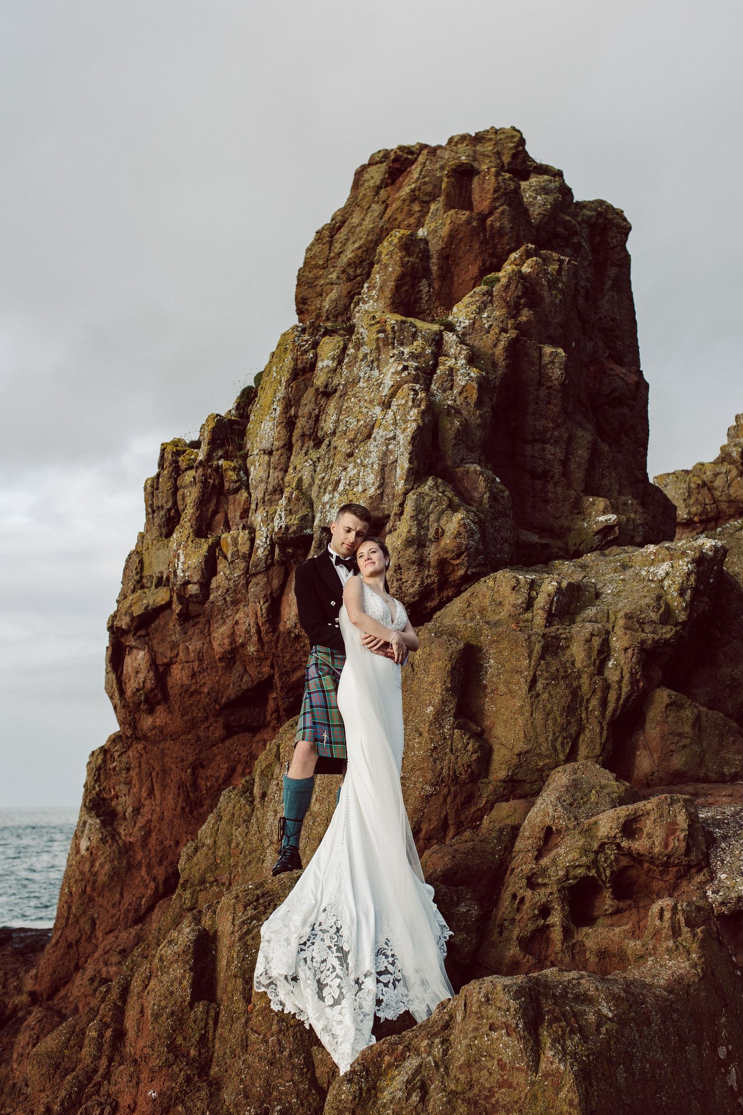 Bride in white dress stands on rocky coastal cliffs while groom watches from below.