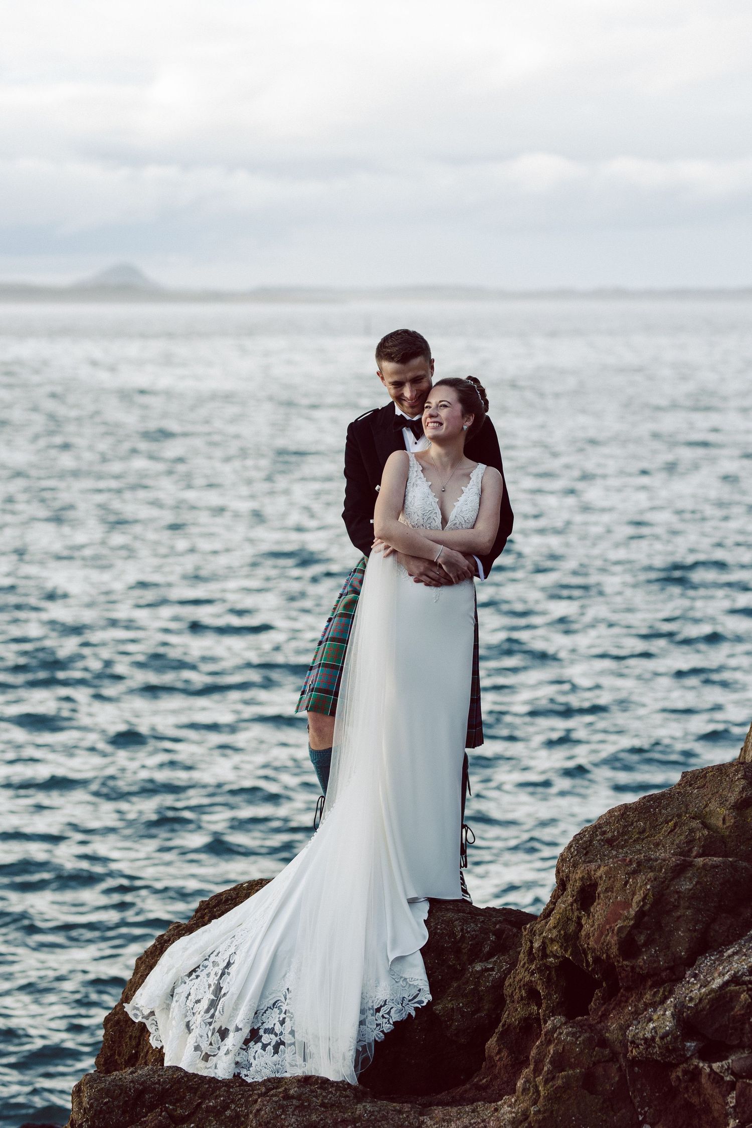 A romantic wedding couple embraces on rocky cliffs overlooking a dramatic ocean landscape at sunset.