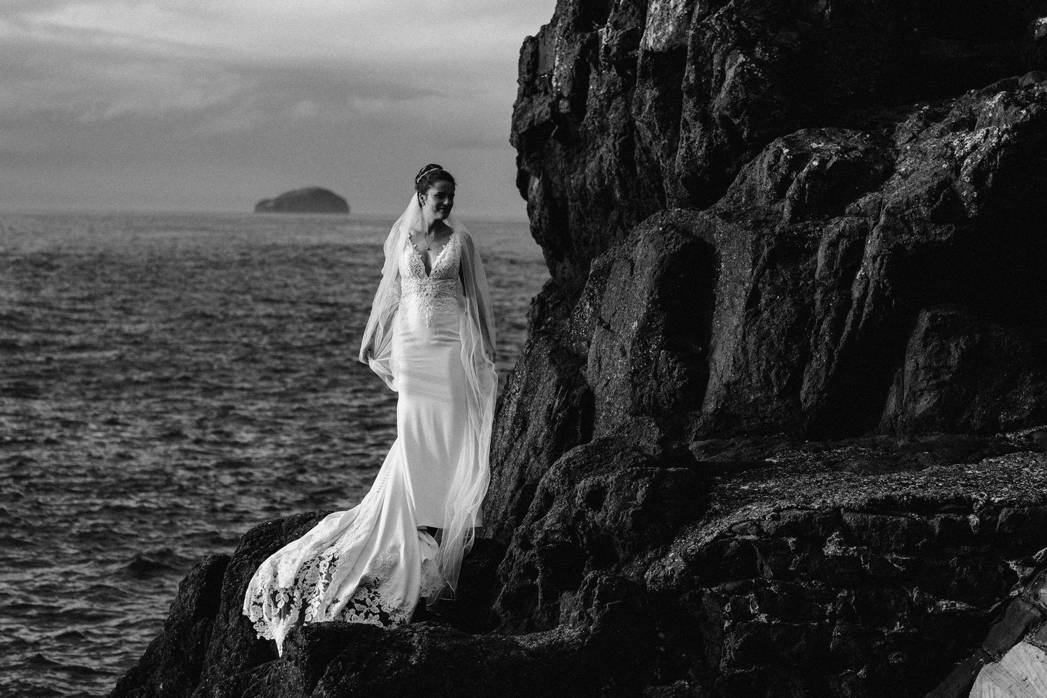 Black and white photo of flowing wedding dress against rocky coastal landscape.