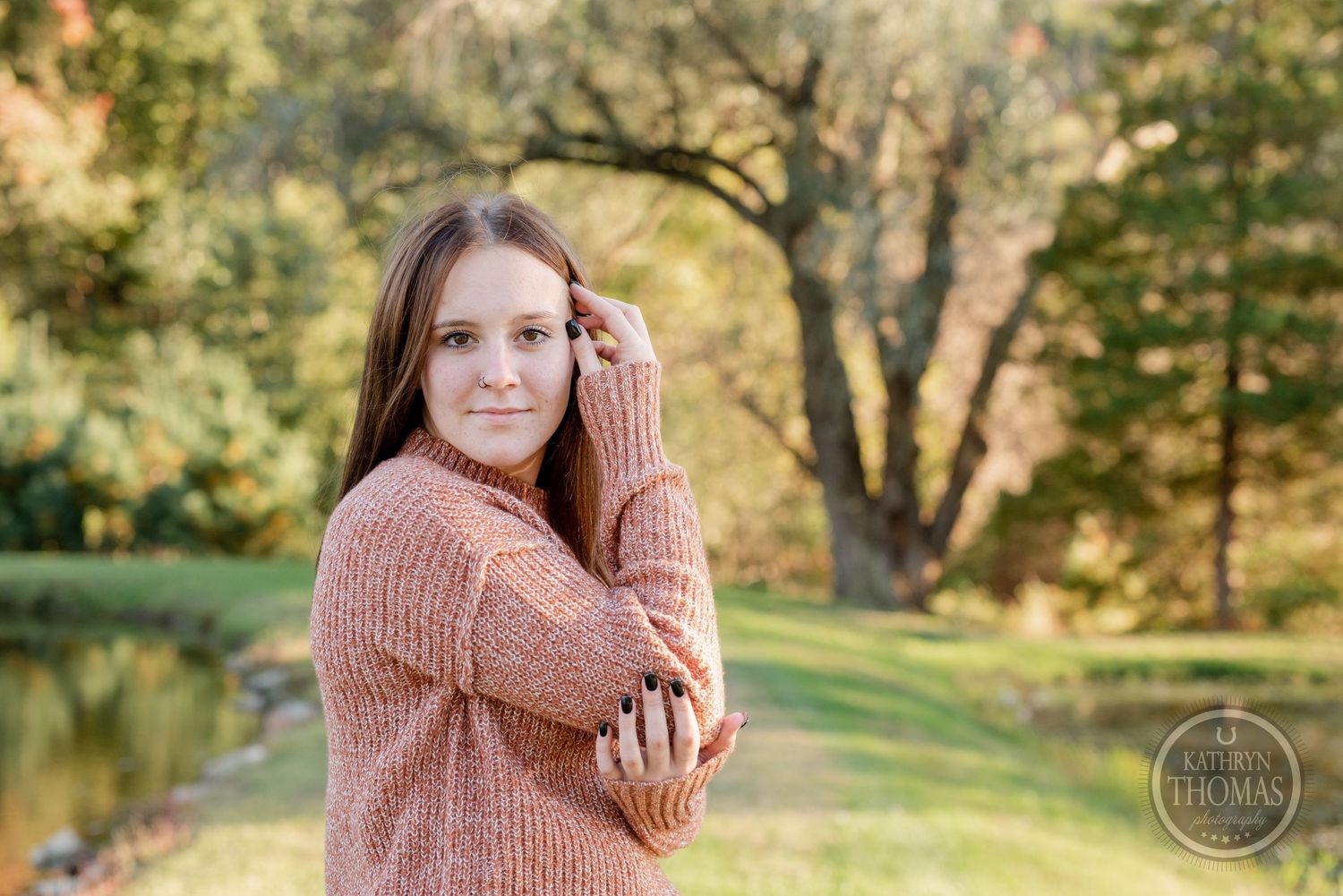 Person in a pink knit sweater poses outdoors in front of autumn trees and a pond on a sunny day.