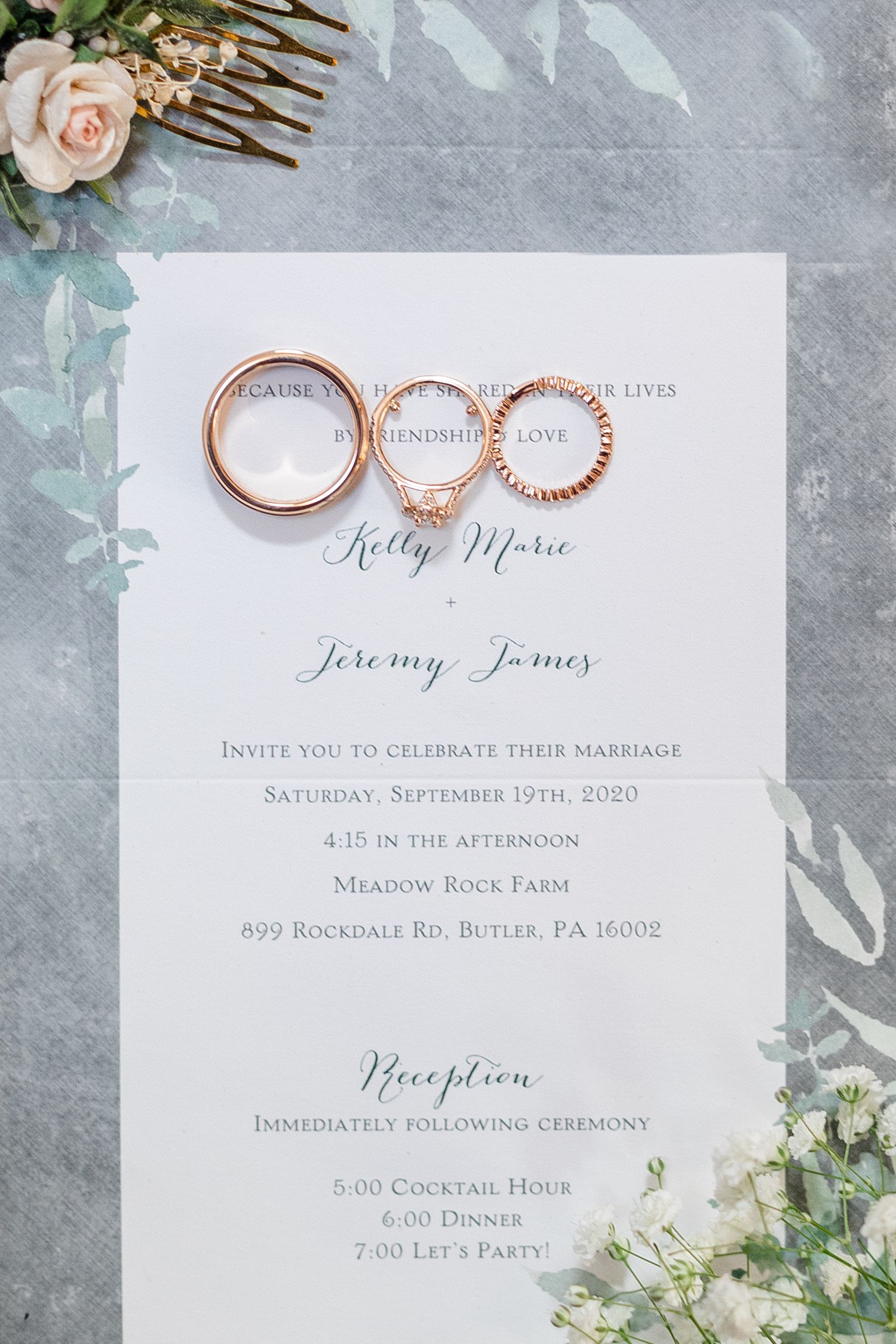 Wedding invitation suite with rose gold rings and floral details on a gray marble background.