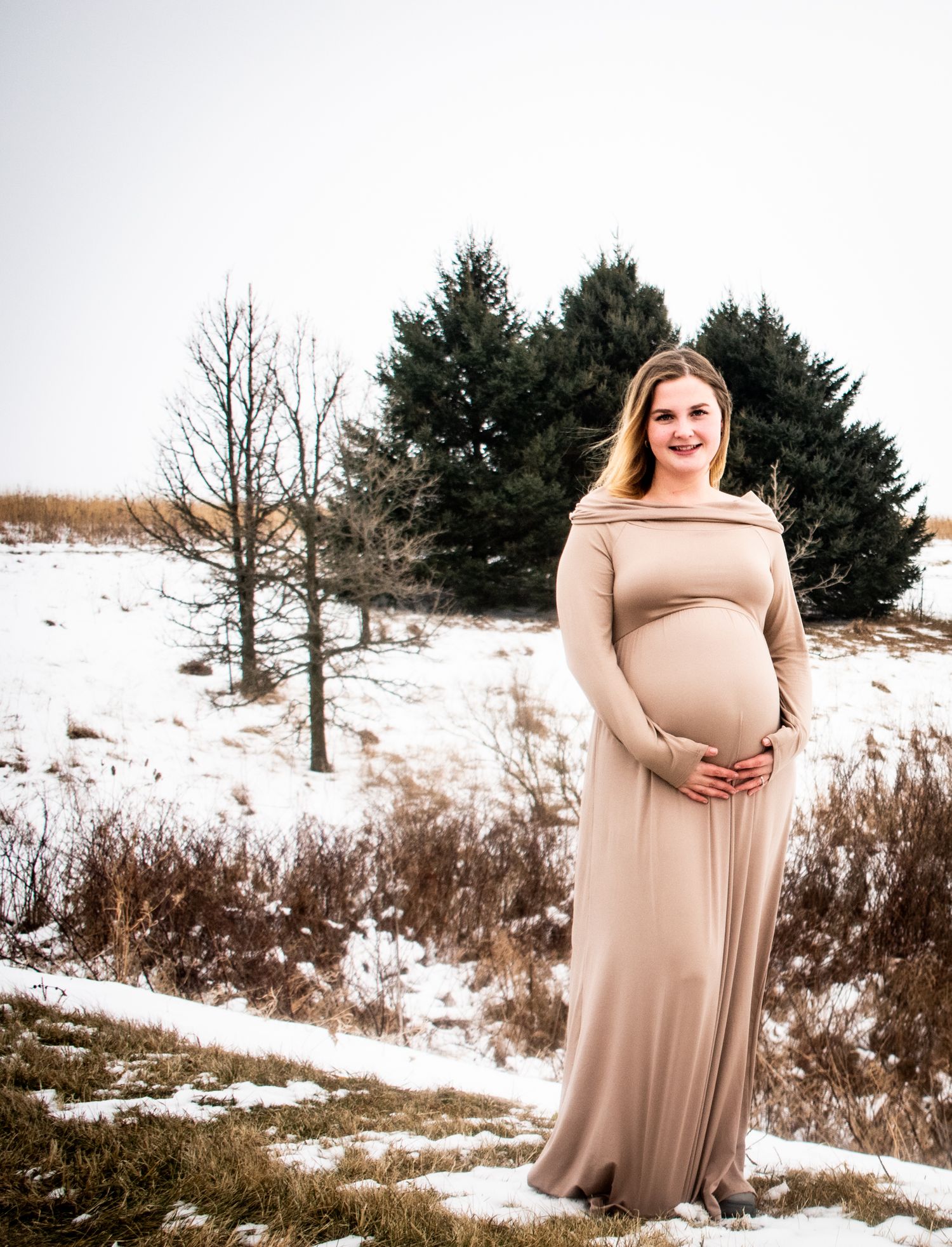A maternity photo of an expectant mother in a beige dress standing in a snowy winter landscape.