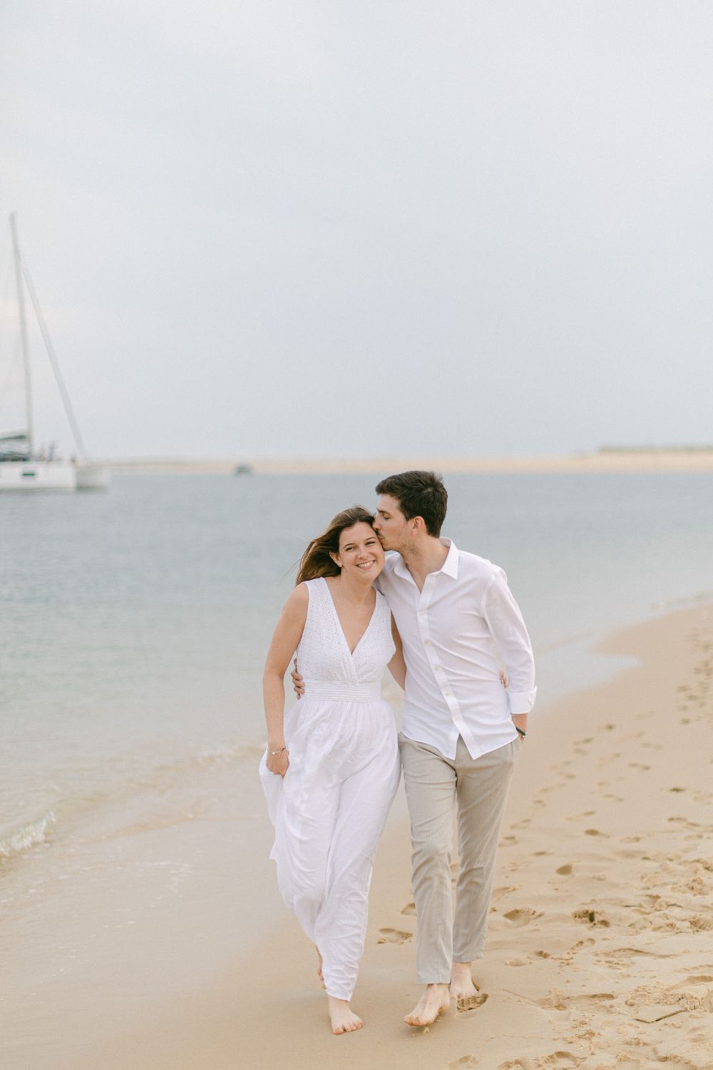 Un couple heureux marchant pieds nus le long de la plage, avec un voilier en arrière-plan.