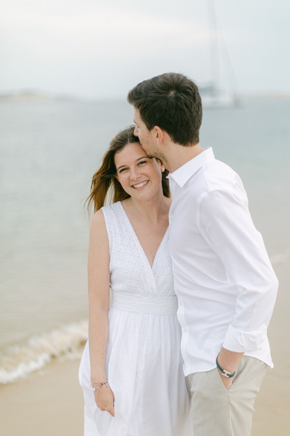 Un couple romantique partageant un moment intime sur une plage brumeuse, l’un portant une robe blanche et l’autre une chemise blanche.