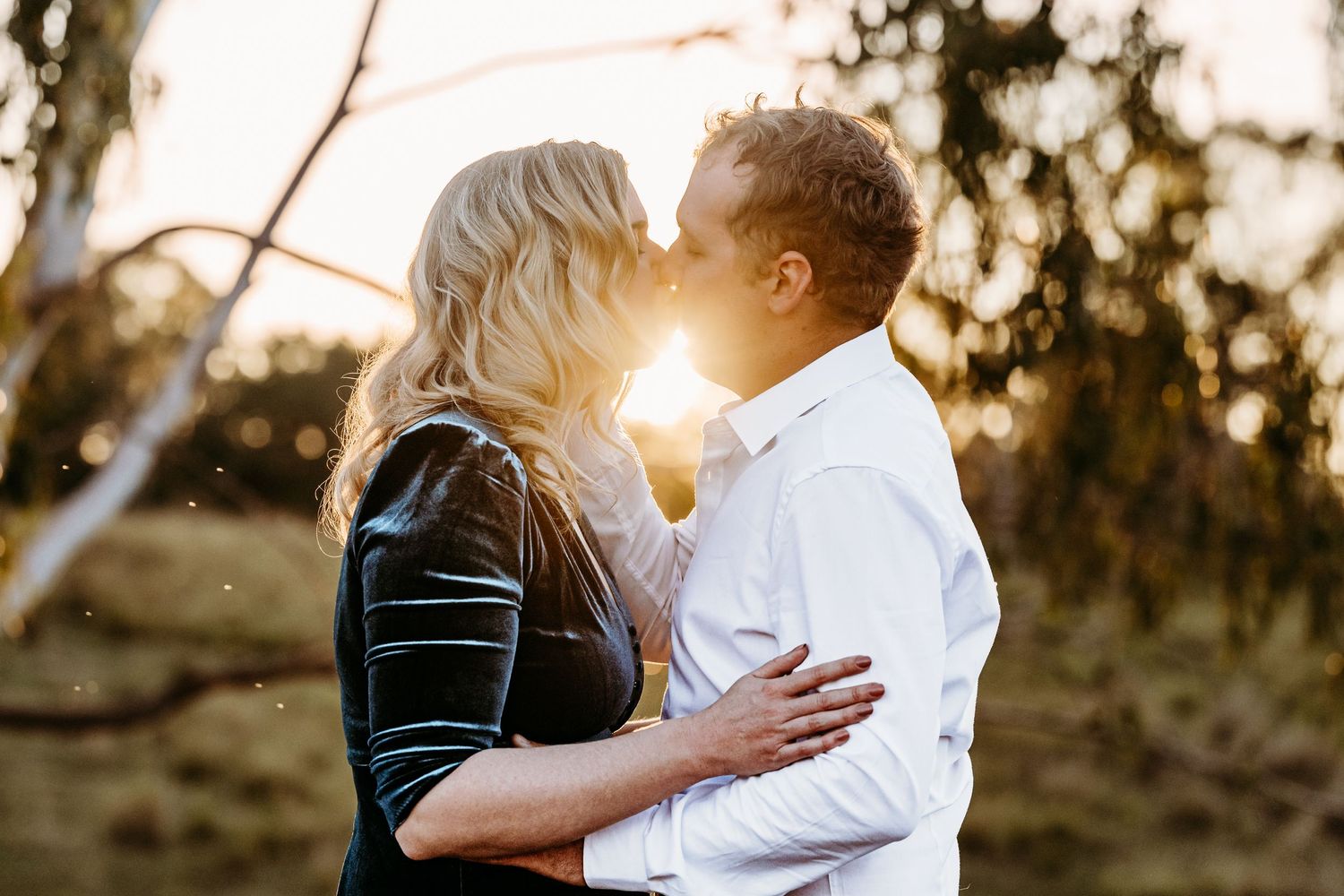 Couple sharing intimate moment in golden sunset light between trees.