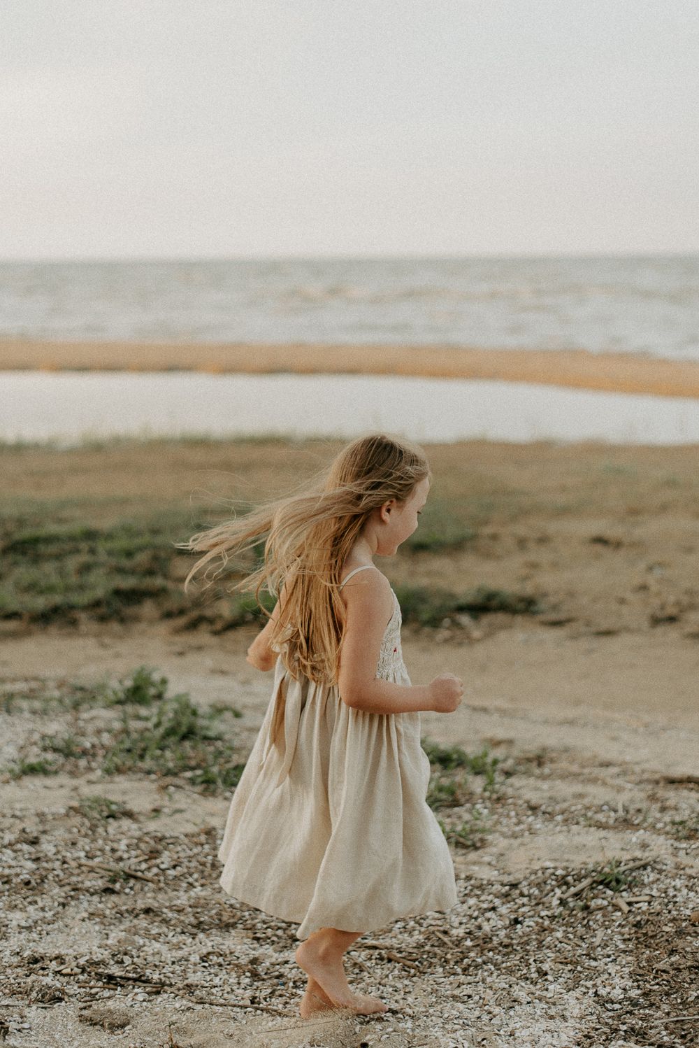 Young girl in white dress twirling on beach with wind-blown hair at sunset.