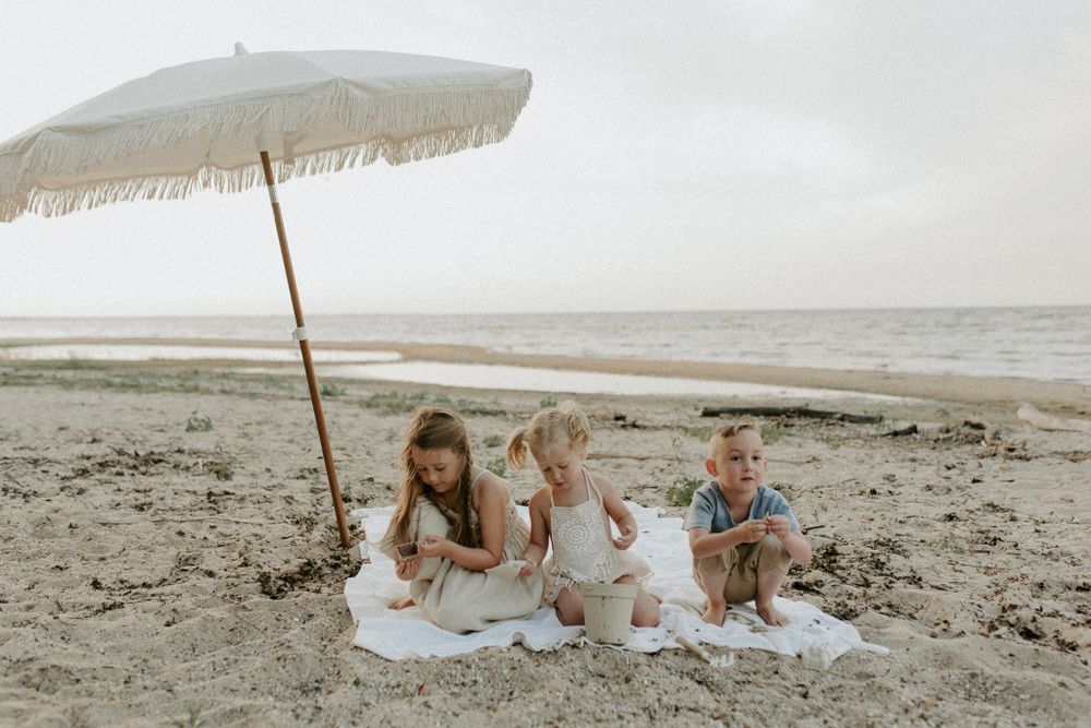 Family picnic on beach under vintage white umbrella on overcast day.