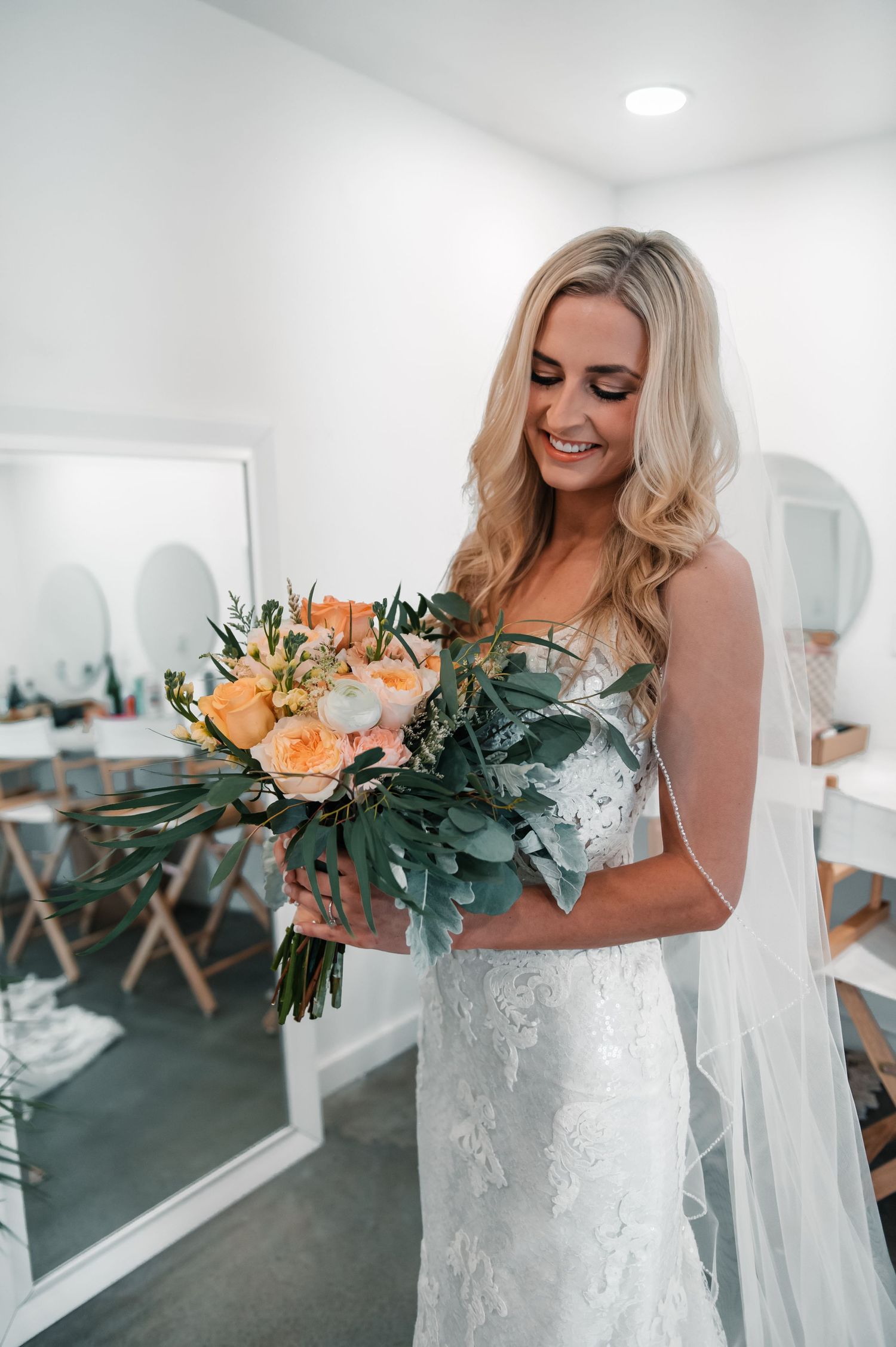 A bridal bouquet with peach roses and greenery held against a white dress.