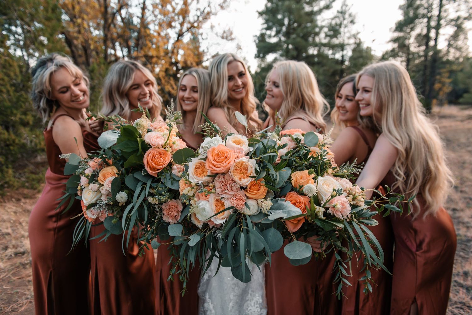 Group of bridesmaids in rust-colored dresses holding floral bouquets outdoors.