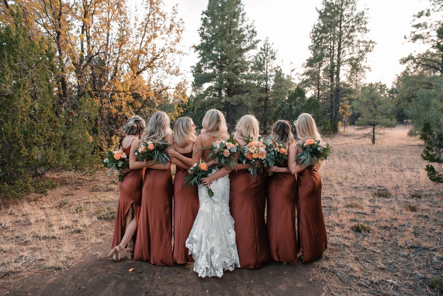 Wedding party in rust-colored attire gathered for group photos in natural outdoor setting.