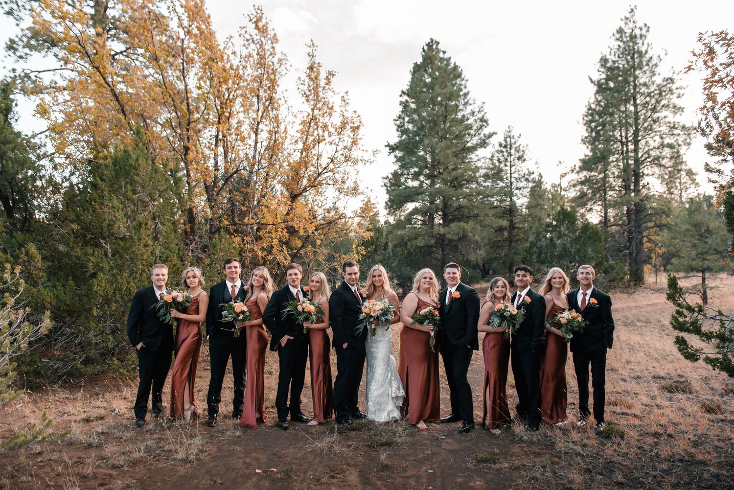 Wedding party members lined up outdoors with pine trees and mountain backdrop.