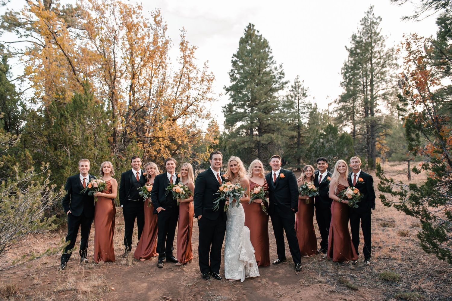 Wedding party poses together on a hillside with autumn trees and evergreens in the background.