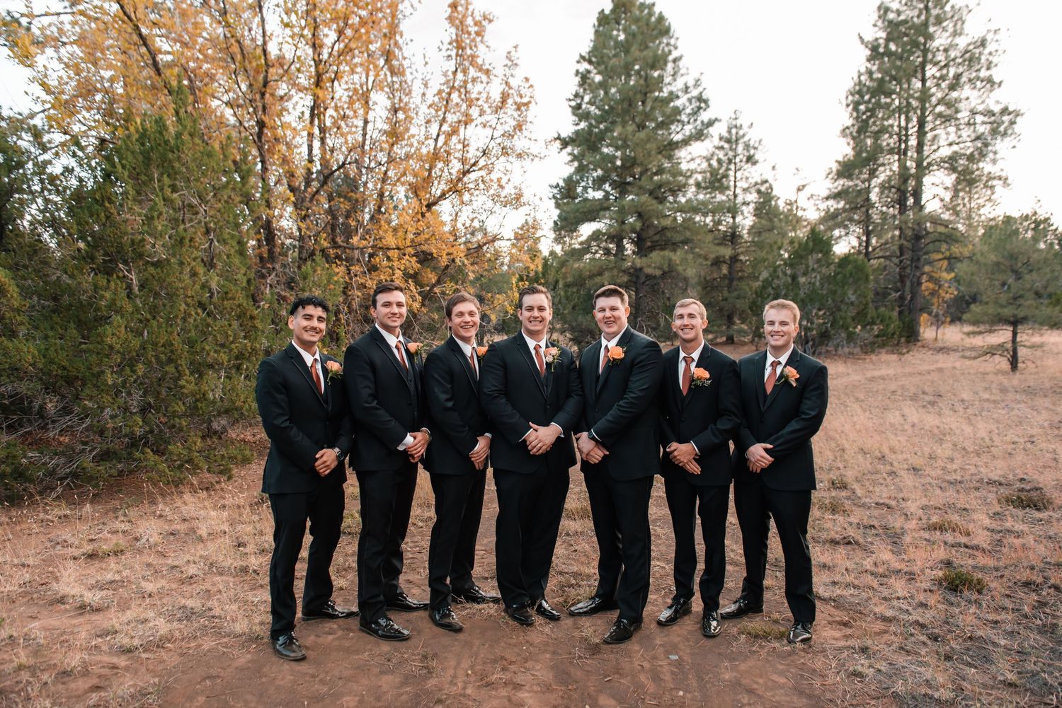 Groomsmen pose together on rustic terrain with pine trees in background.