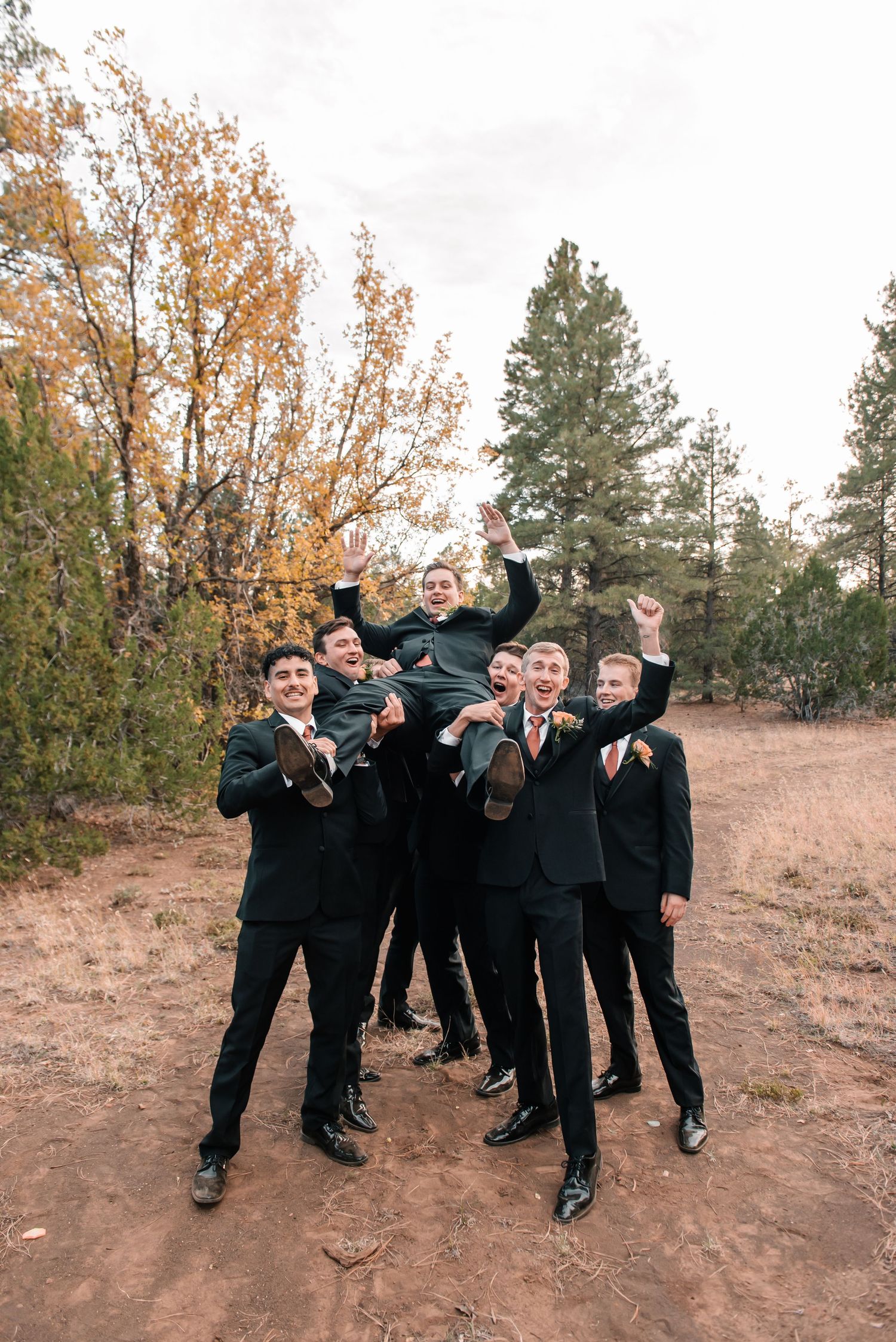 Groomsmen in black suits jump playfully for group photo on hillside.