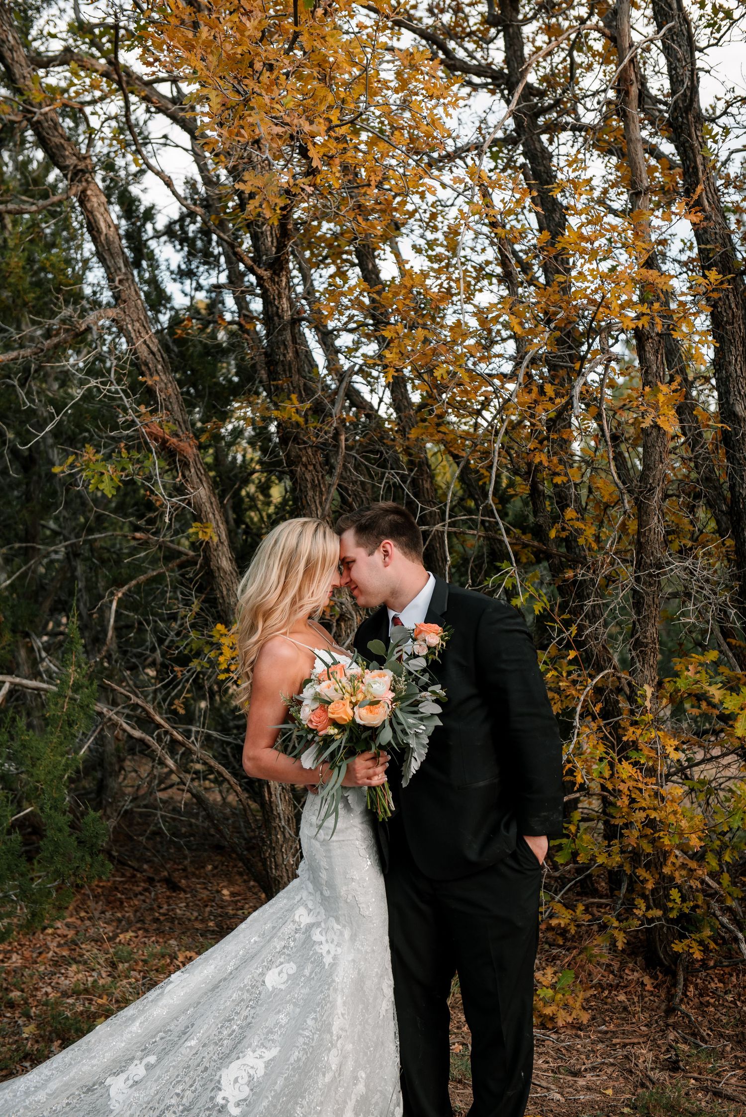 Romantic wedding portrait beneath autumn trees with peach bouquet.