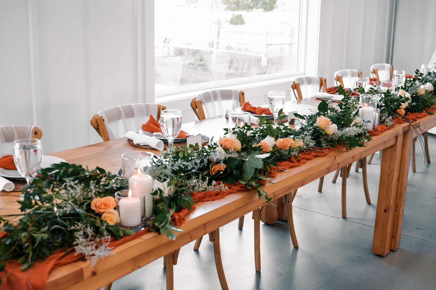 Long wooden tables decorated with greenery garlands and candles in a bright white venue.