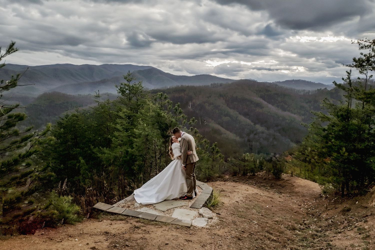 White wedding dress billows in mountain breeze against dramatic cloudy mountain vista.