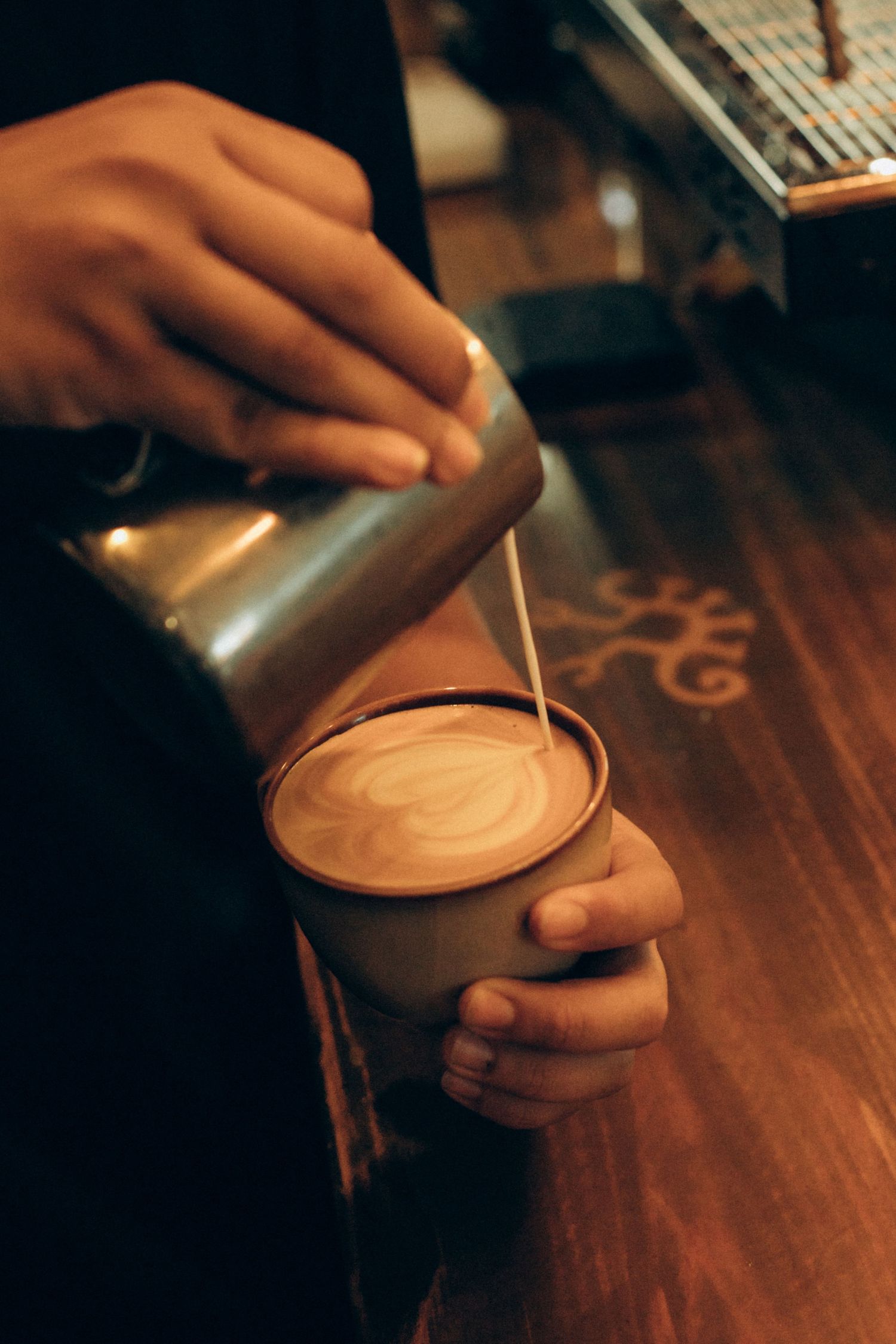 Hands pouring steamed milk into coffee creating latte art on wooden counter.