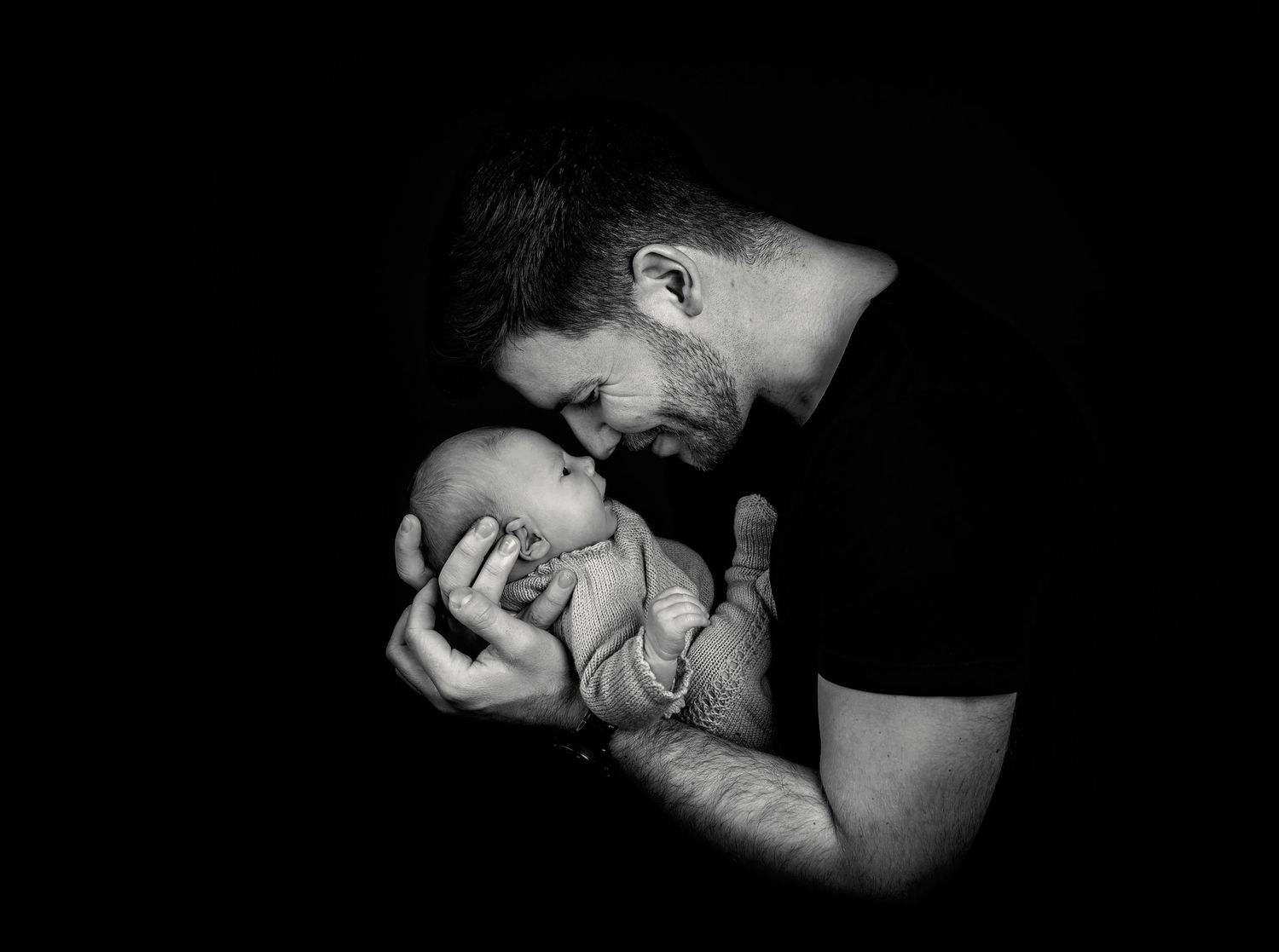 Tender black and white portrait of parent cradling newborn baby against dark background.