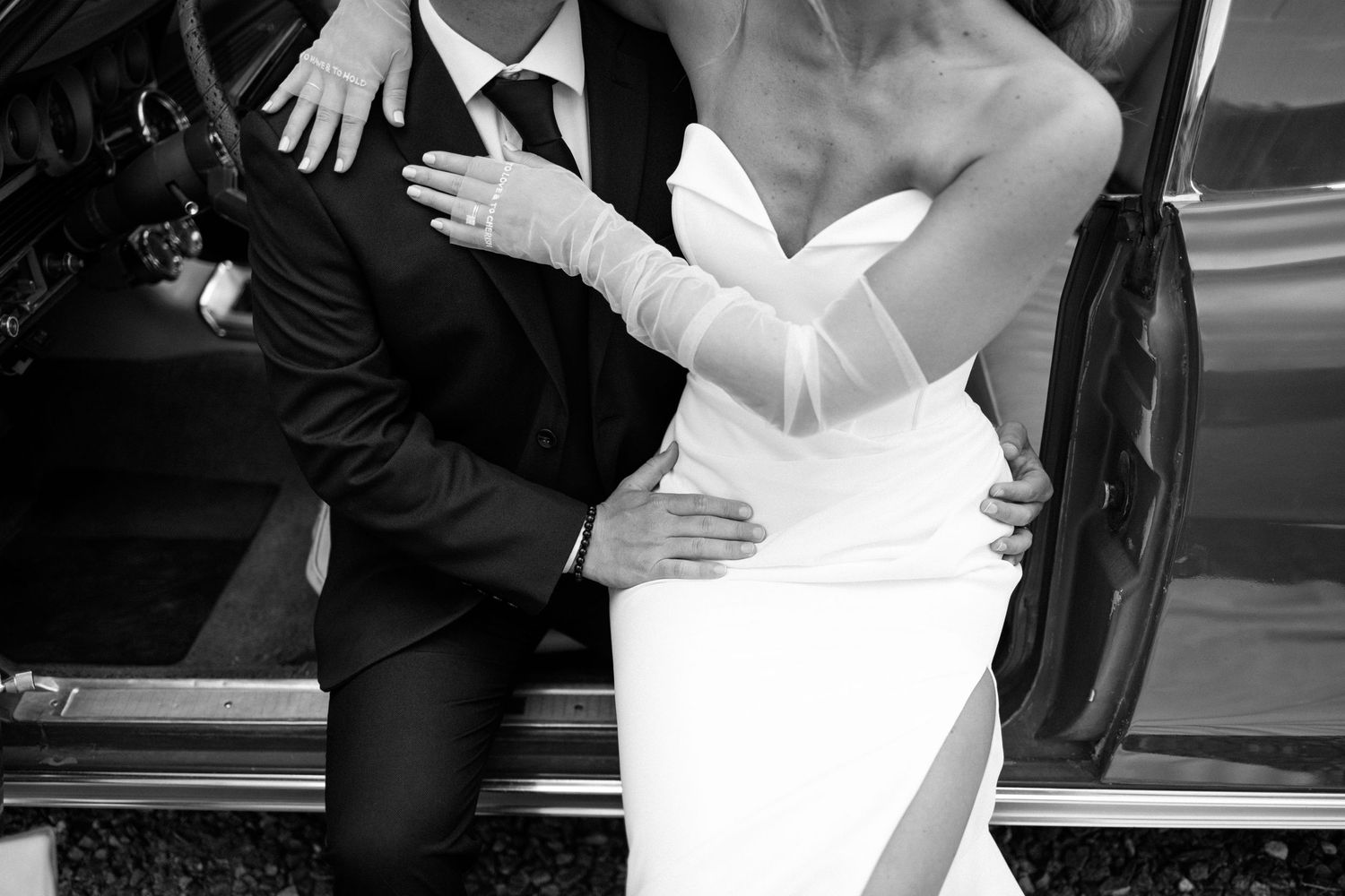 Black and white candid of newlyweds embracing by vintage car.