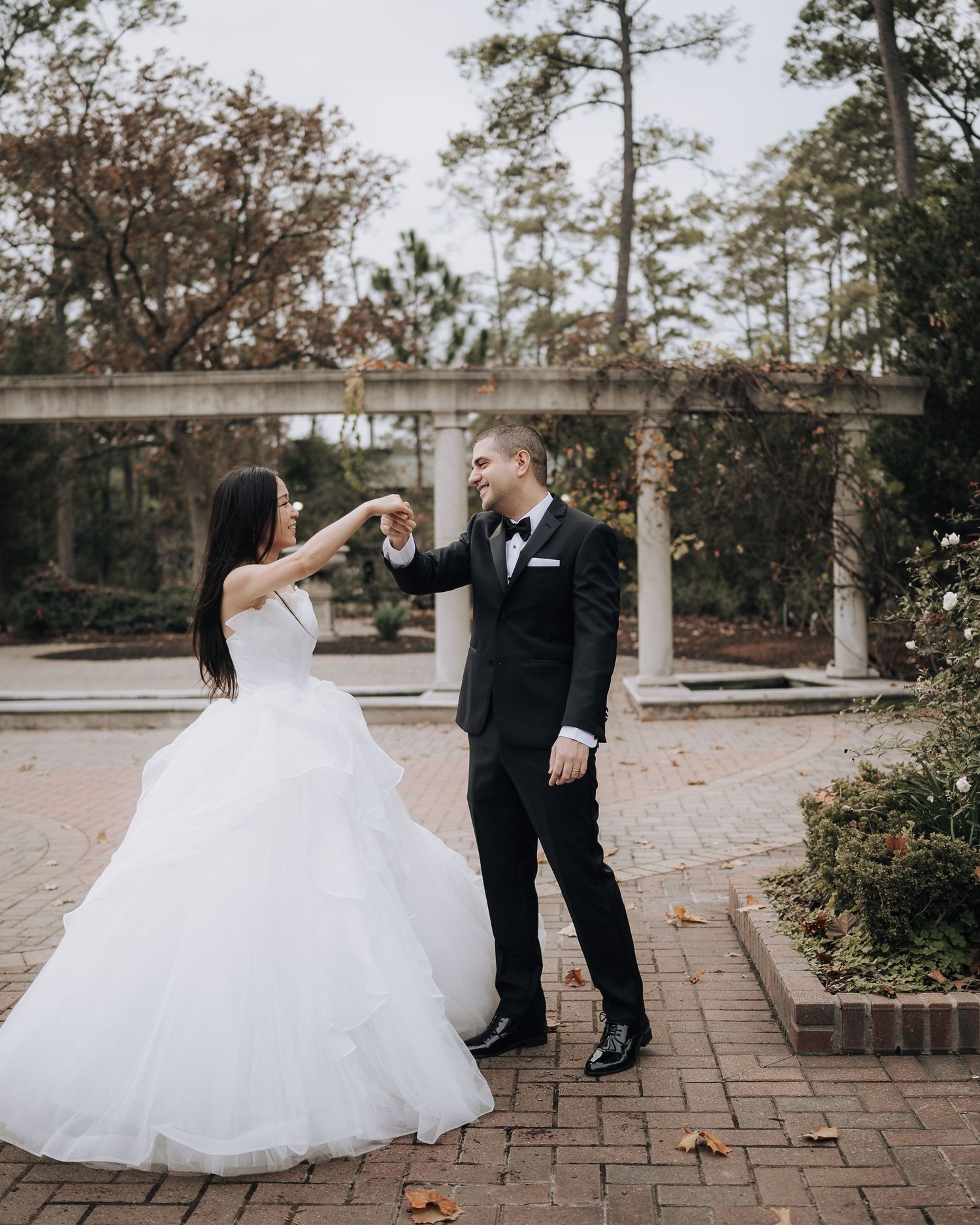Playful wedding couple dancing and twirling on a brick pathway surrounded by trees.